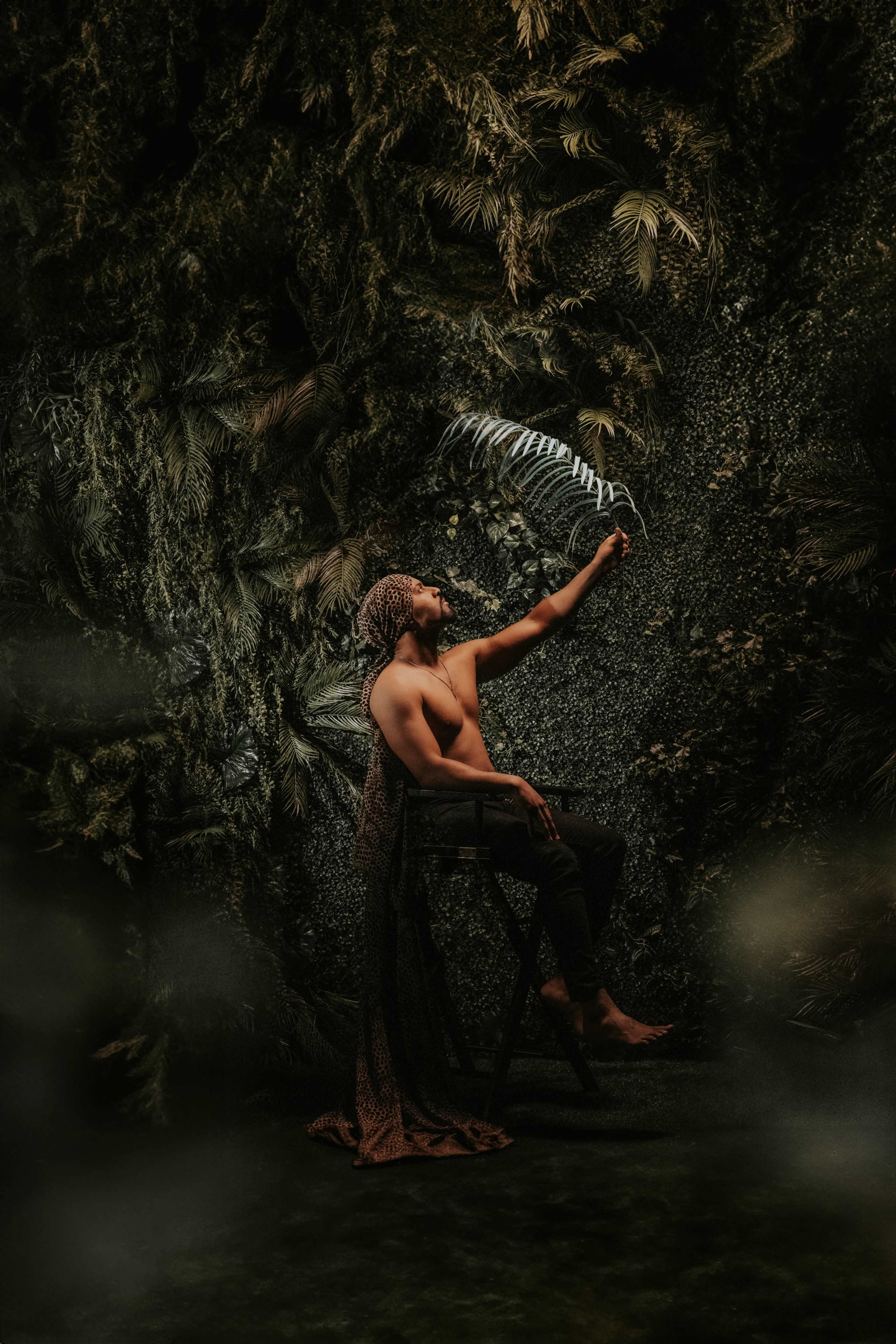 A man with a scarf draped over his shoulders sits on a stool against a backdrop of lush greenery, reaching out to a fern.