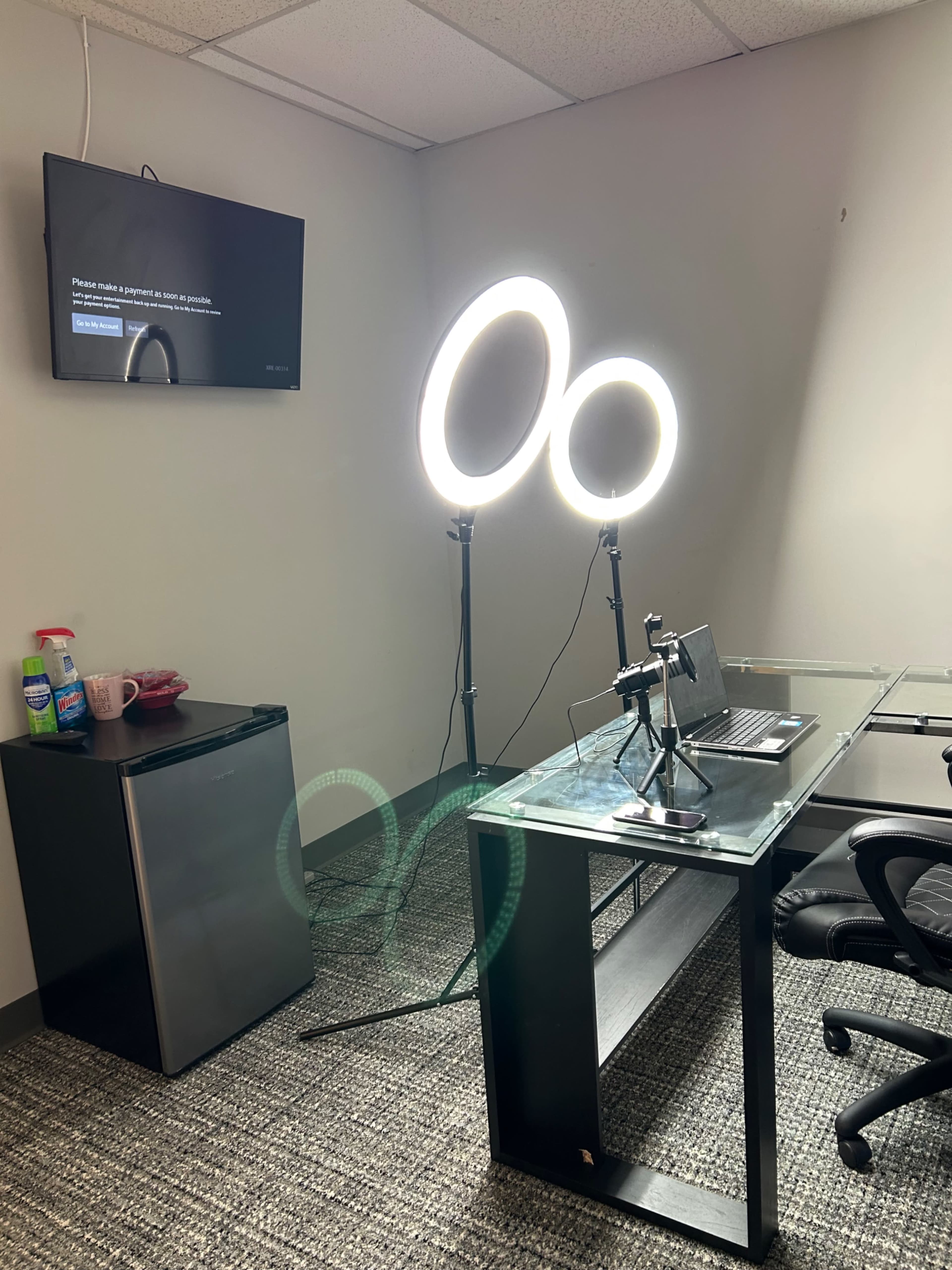 A small office space with a desk, a chair, a laptop, two ring lights on stands, a TV mounted on the wall, and a mini refrigerator in the corner.