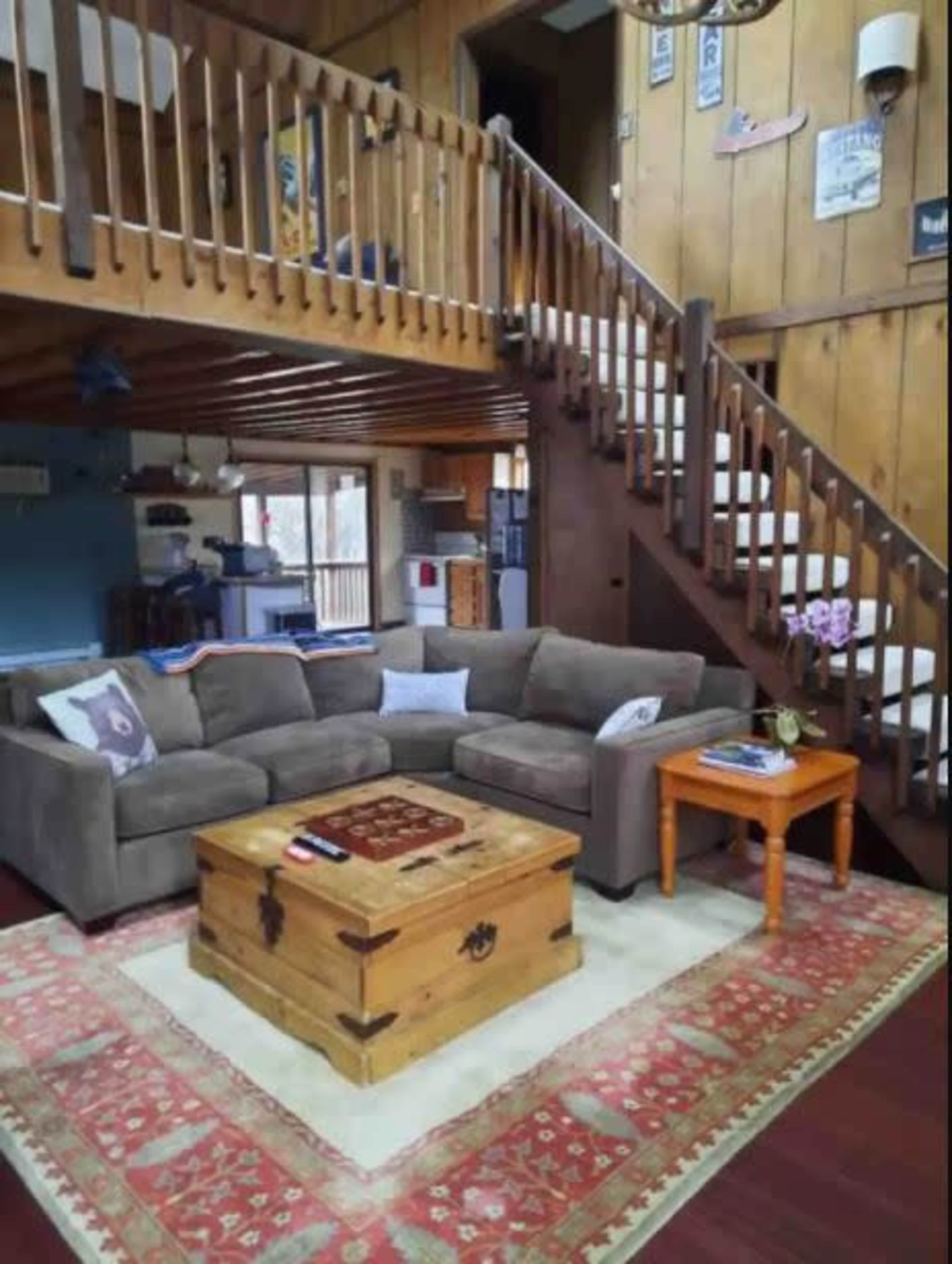 The image shows a cozy living room with a gray sectional sofa, a wooden coffee table, and a staircase leading to an upper level.
