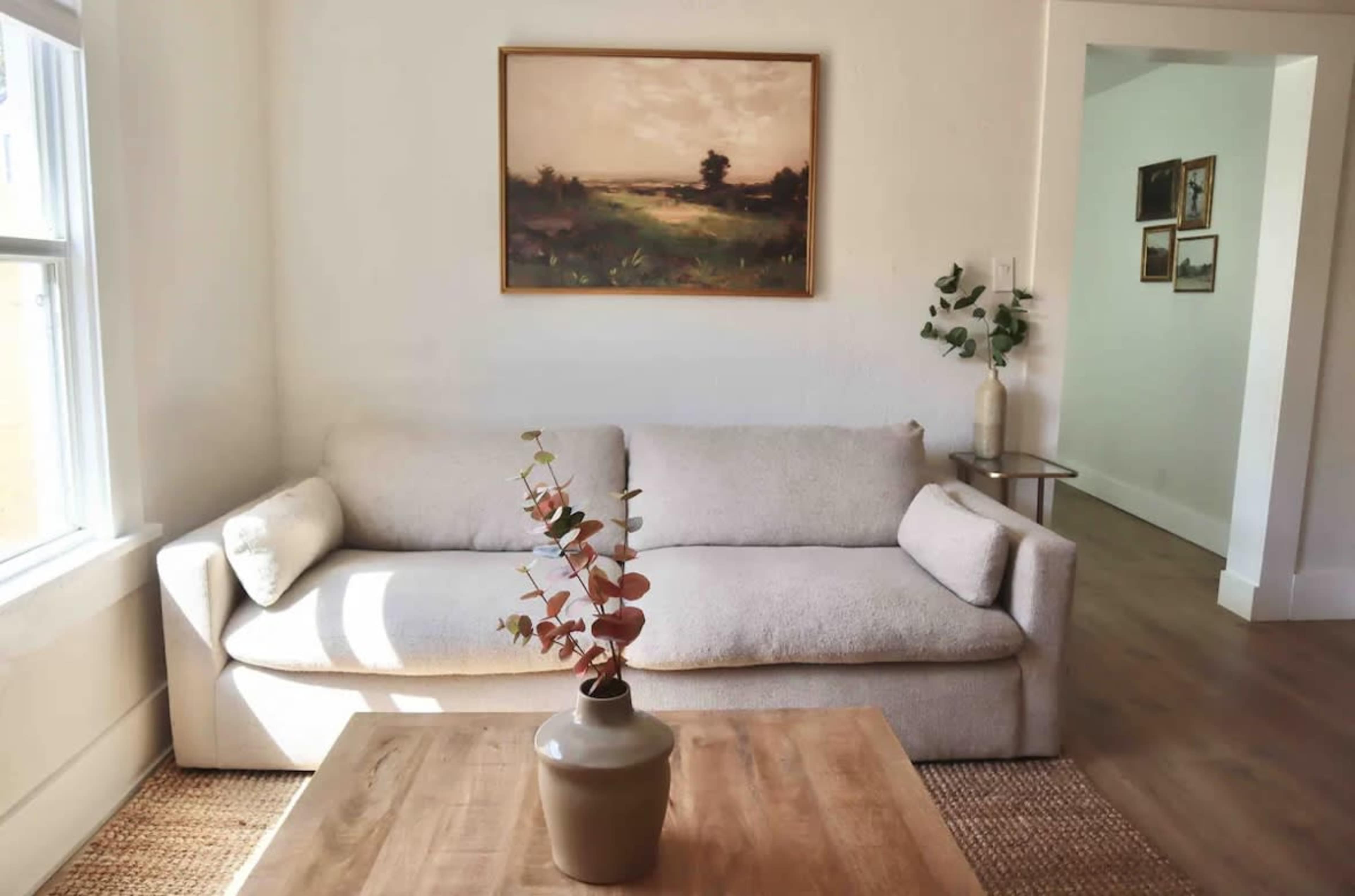 A light-filled living room features a beige sofa with a simple vase and a small branch on a wooden coffee table, framed by a painting on the wall and a doorway leading to another room.