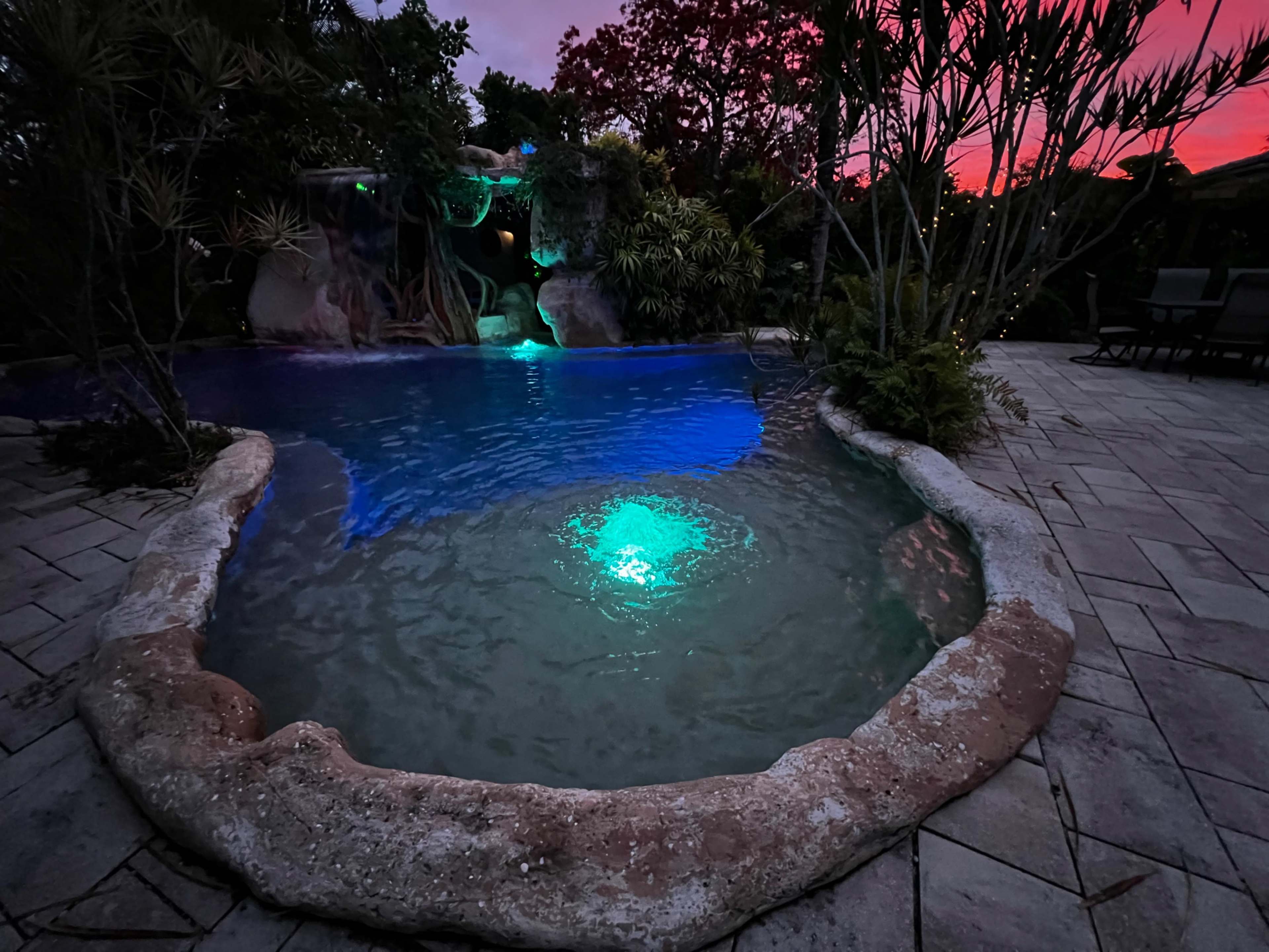 The image shows a pool with a glowing blue light, surrounded by natural rock features and tropical vegetation, under a colorful sunset sky.