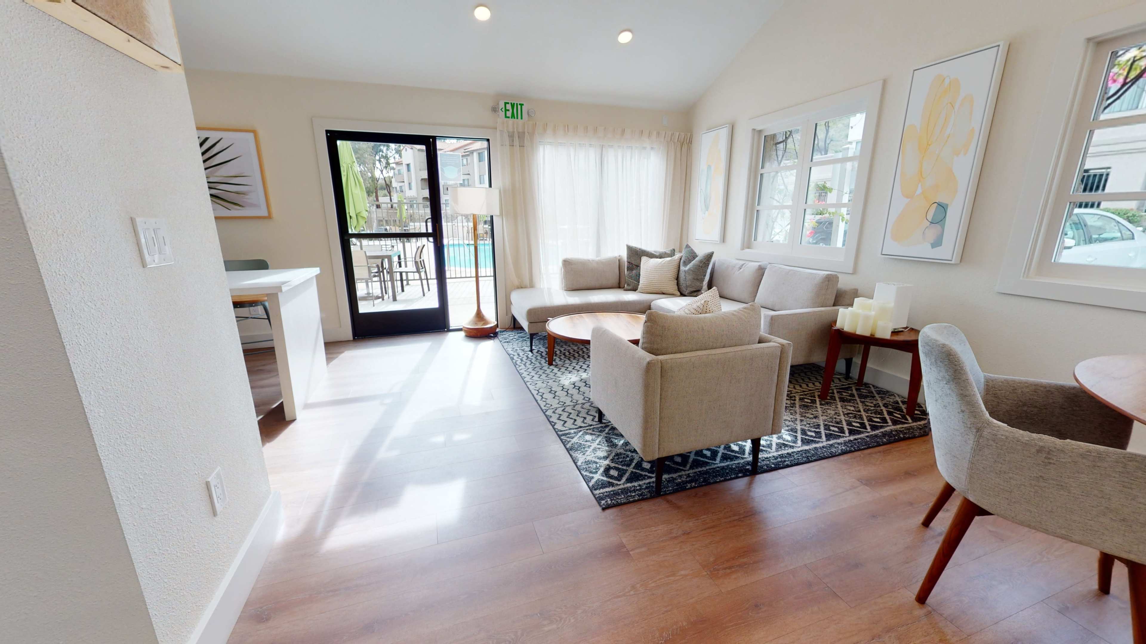 A sunlit living room featuring a beige couch, matching chairs, a wooden table, and large windows overlooking a pool area.