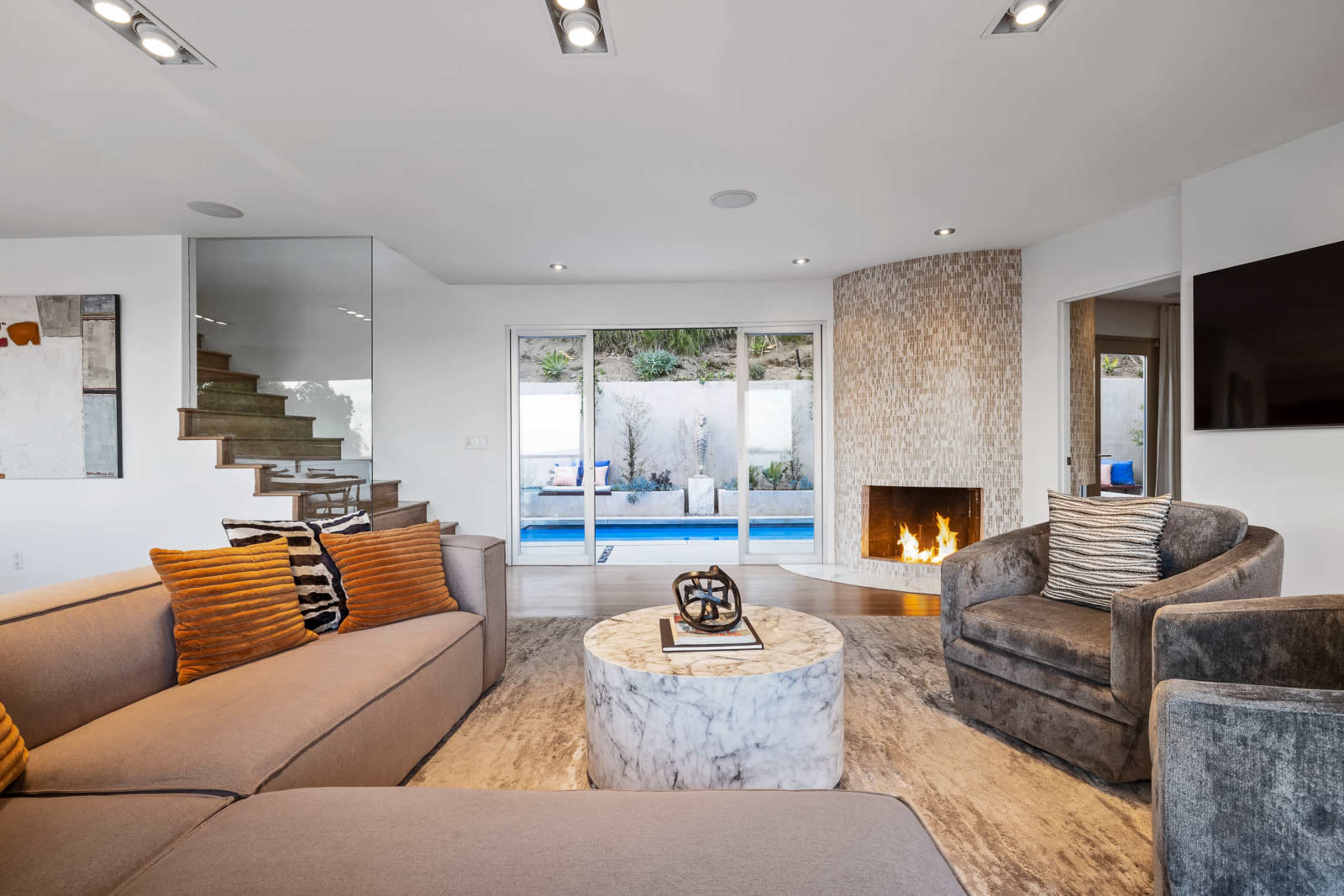 The image shows a modern living room featuring a marble coffee table, a sectional gray sofa with striped cushions, and a cozy fireplace, overlooking a pool area through glass doors.