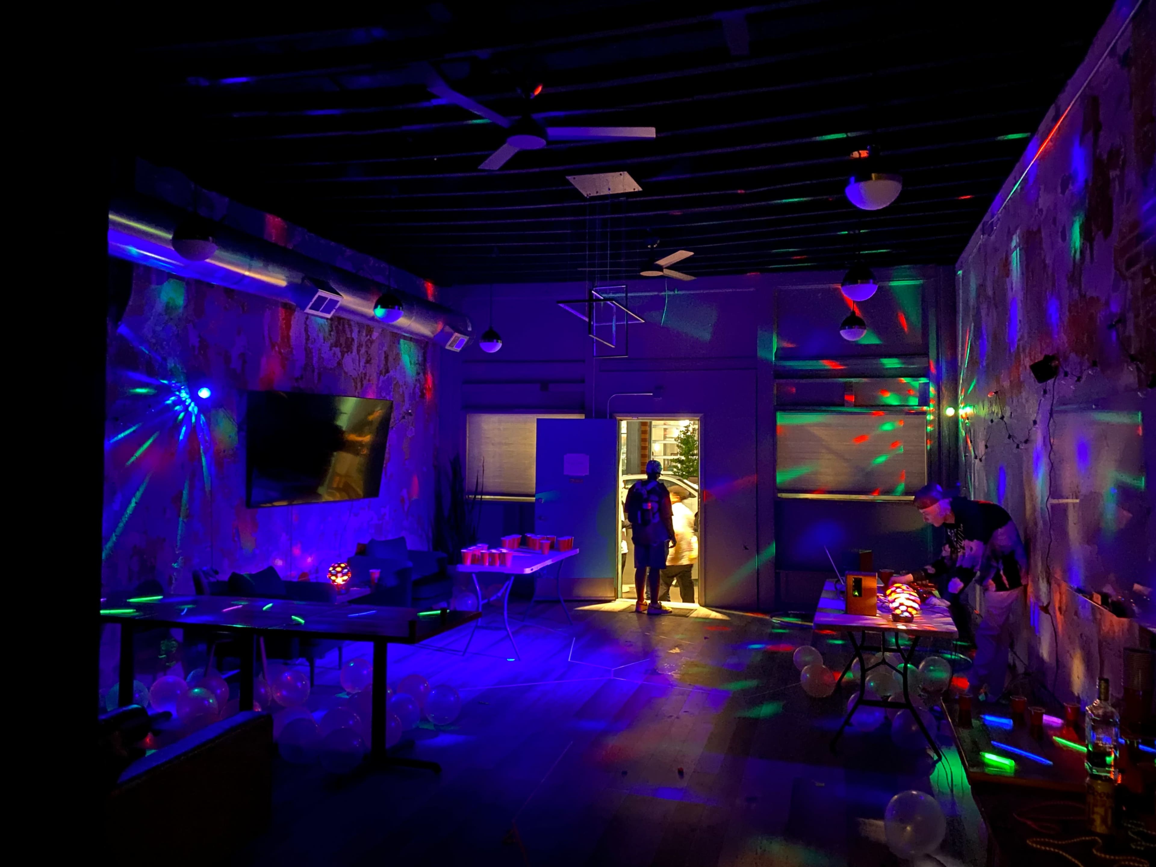 A dimly lit room features colorful lights reflecting off the walls, with party decorations and balloons scattered across the floor.