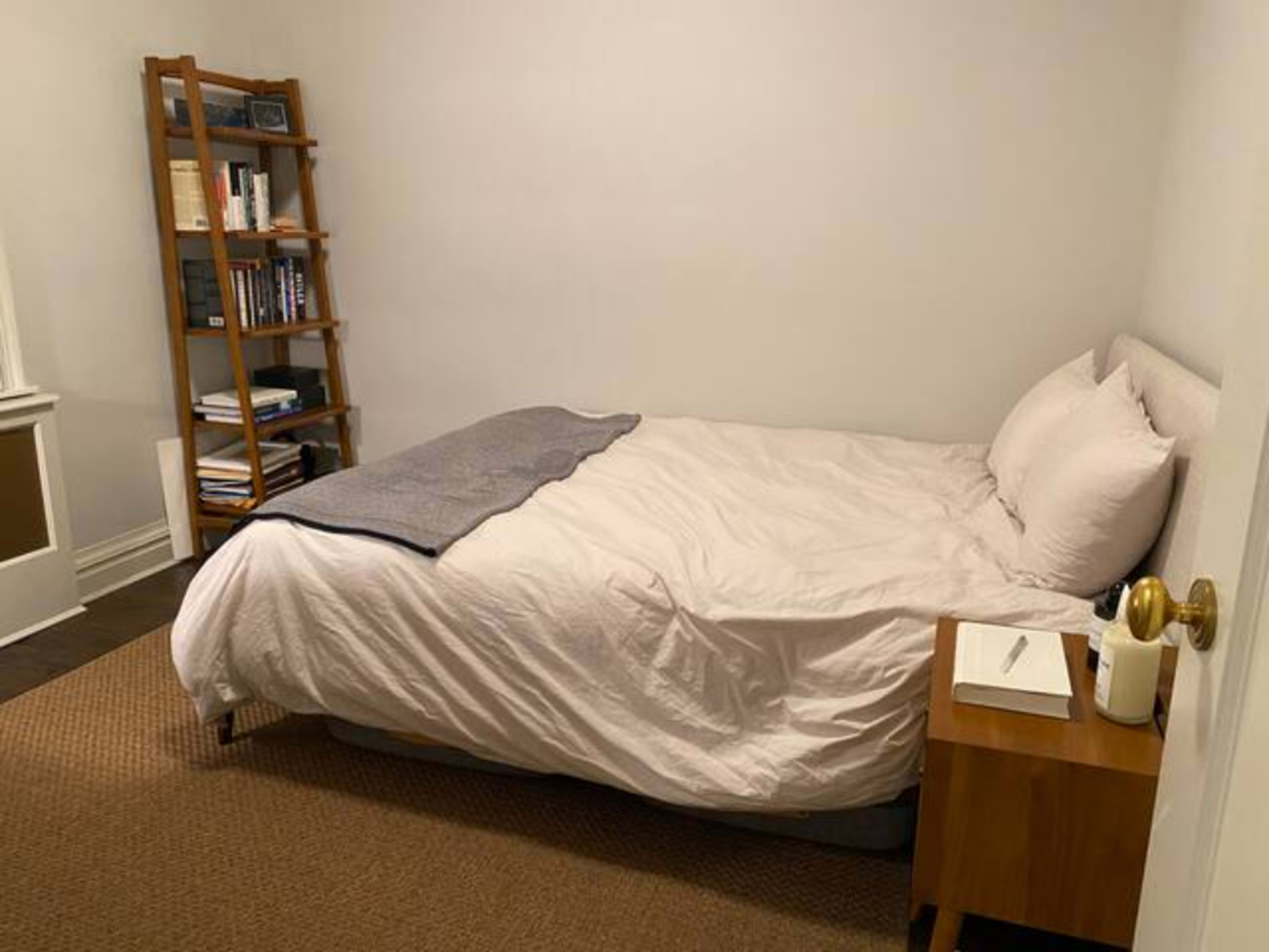 A simple bedroom with a bed, a nightstand, and a bookshelf against a plain wall.