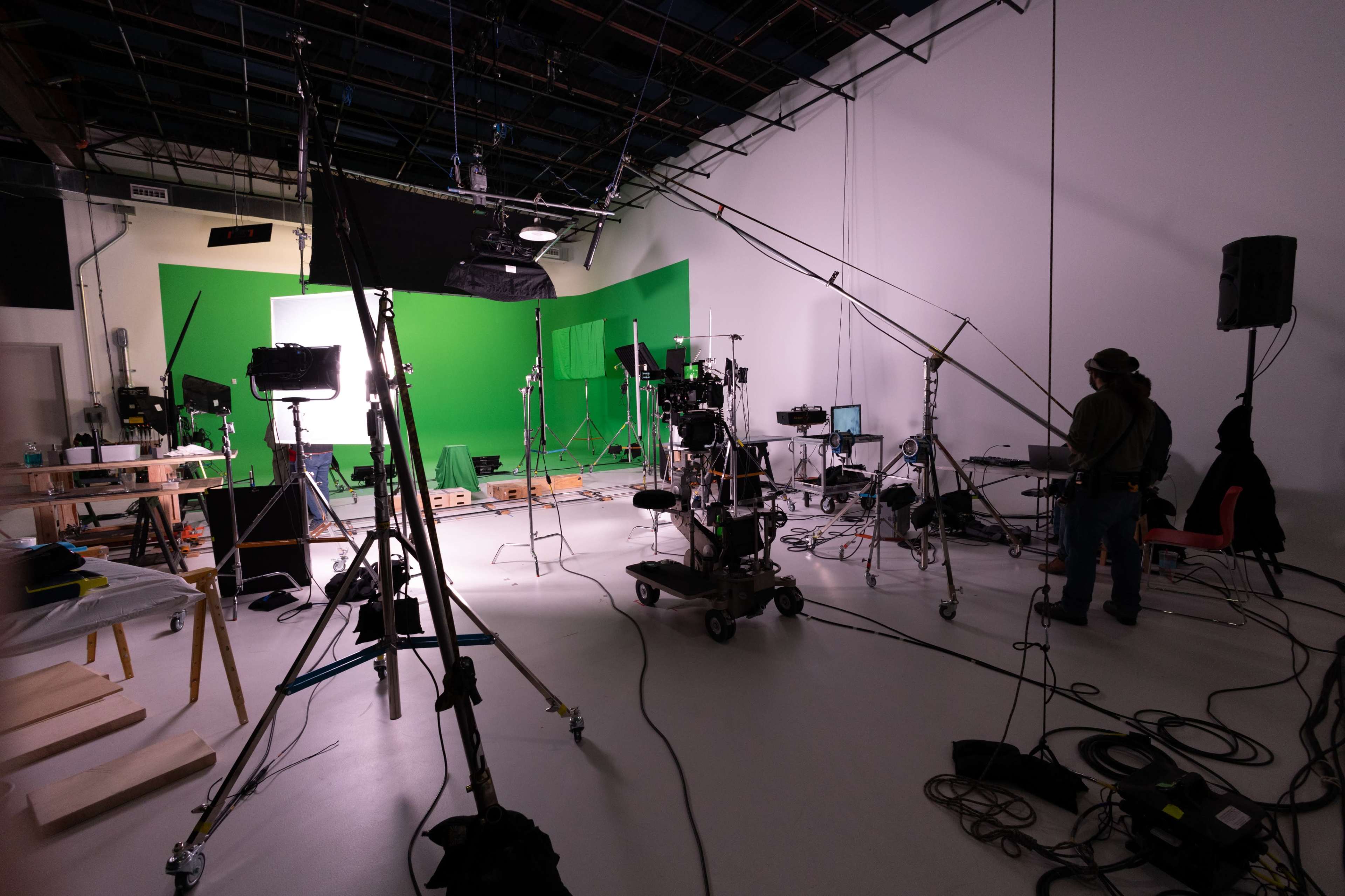 The image shows a film studio set up with multiple cameras, lighting equipment, and a green screen, along with a crew member operating a camera.