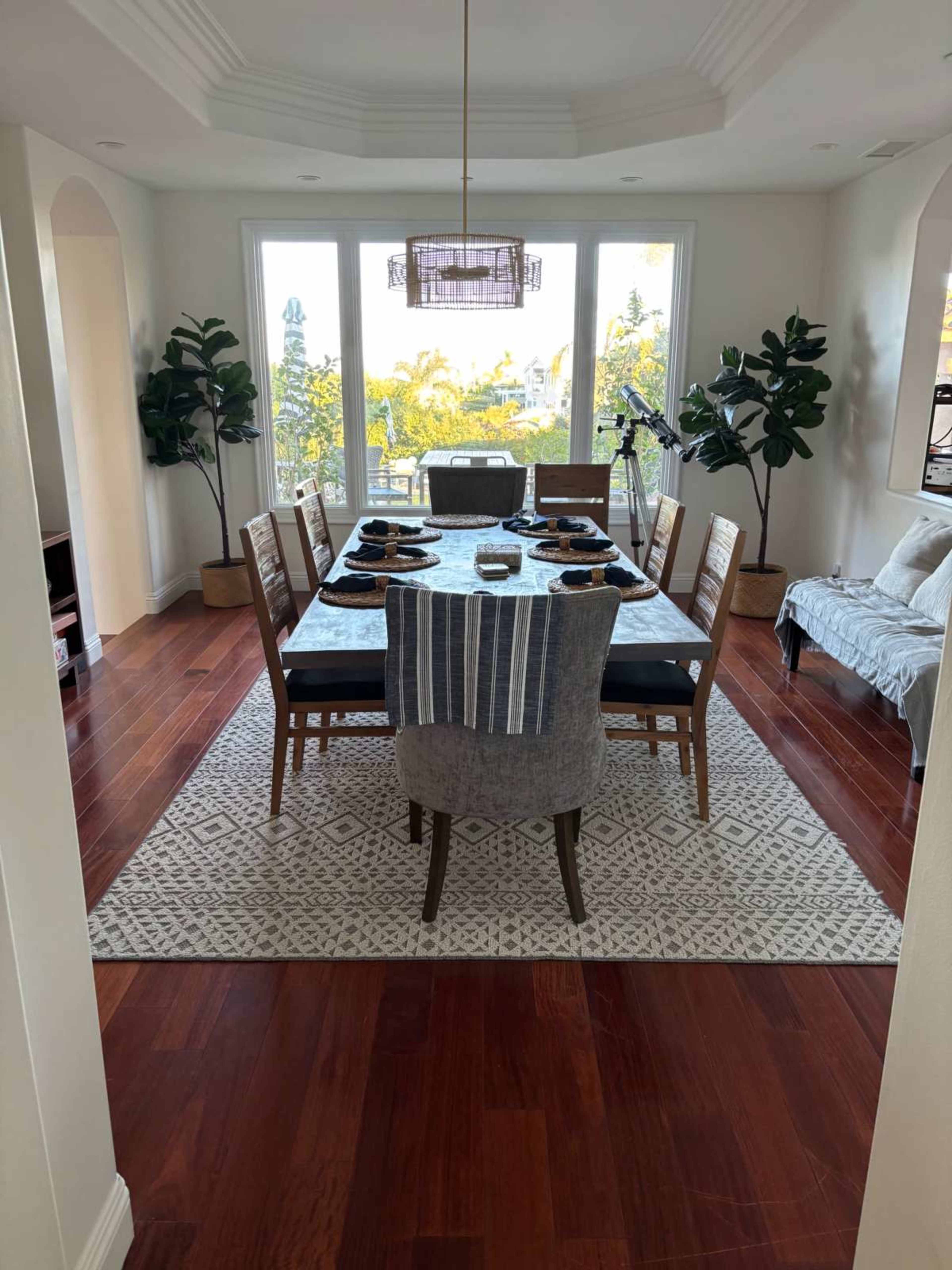 The image shows a dining room with a large table set for meals, surrounded by chairs, and featuring large windows that let in natural light, along with decorative plants and a rug on a hardwood floor.