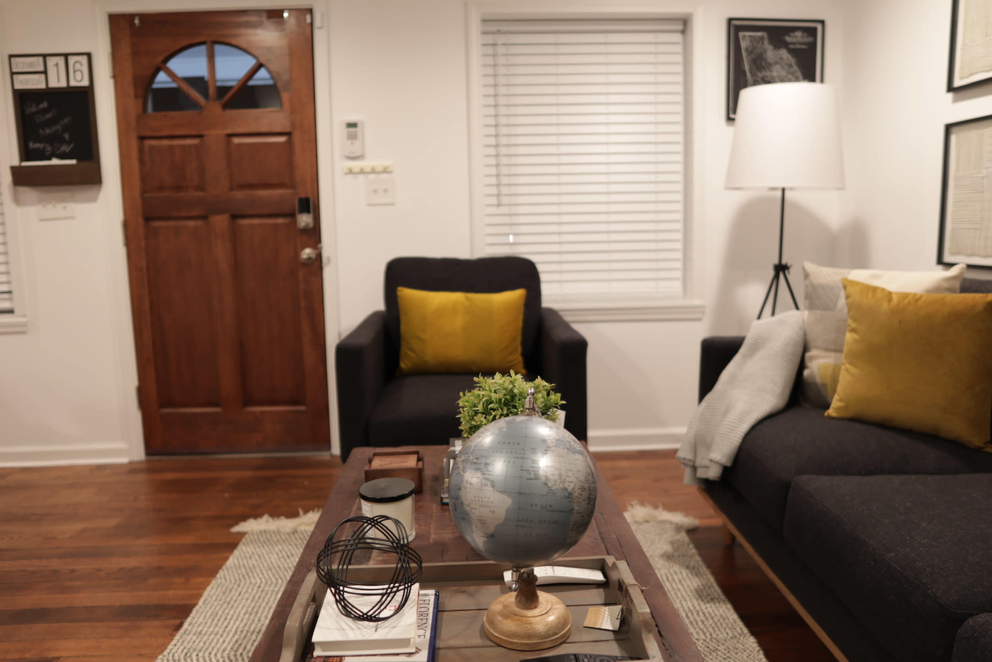 A cozy living room with a wooden front door, two chairs, a coffee table featuring a globe, and a lamp in the corner.
