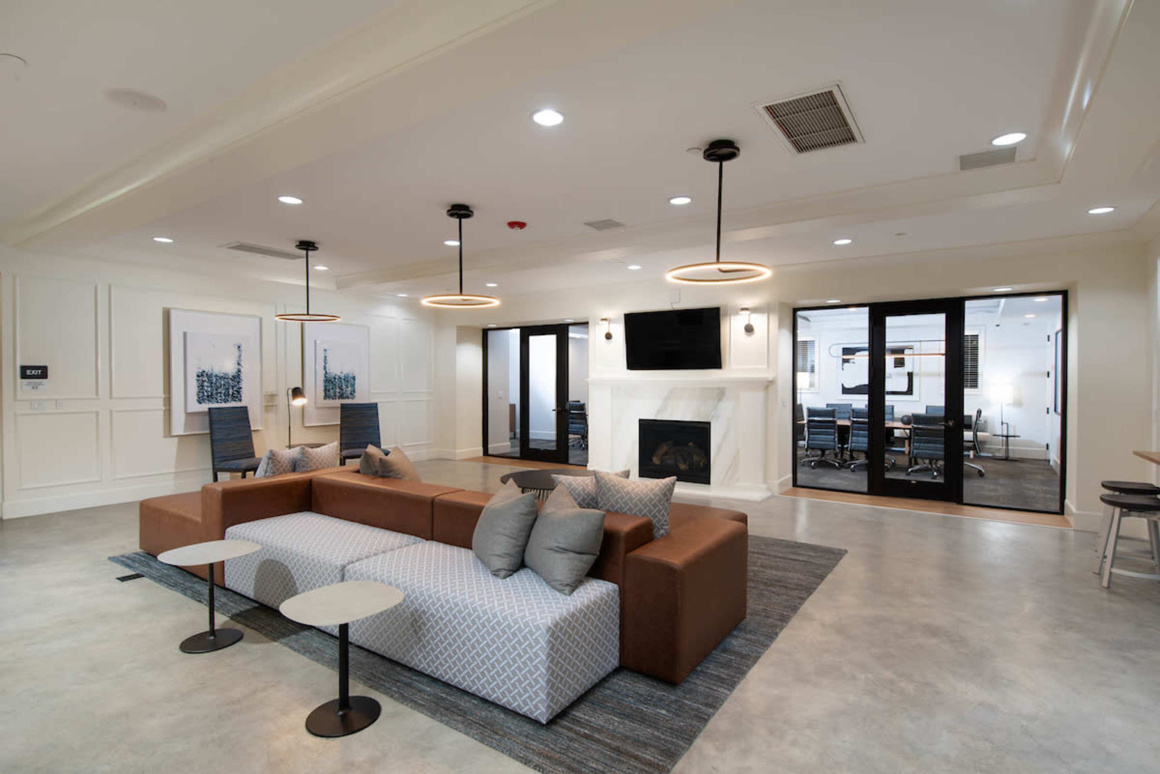 A modern communal lounge features a sectional sofa, side tables, pendant lights, and a fireplace, with glass doors leading to a conference room.