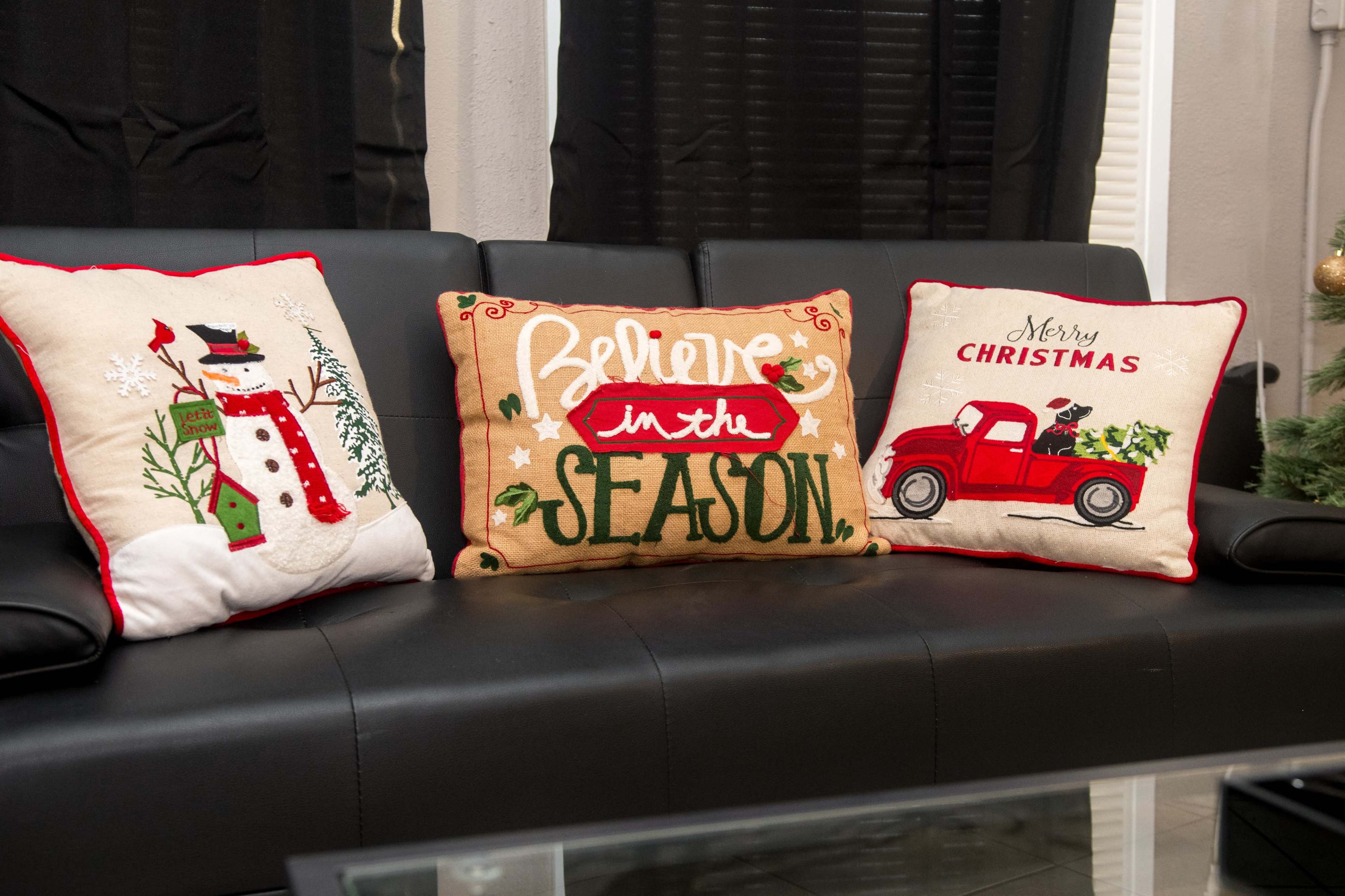 Three decorative pillows with holiday themes are arranged on a black sofa, showcasing a snowman, the phrase "Believe in the Season," and a red truck with Christmas trees.