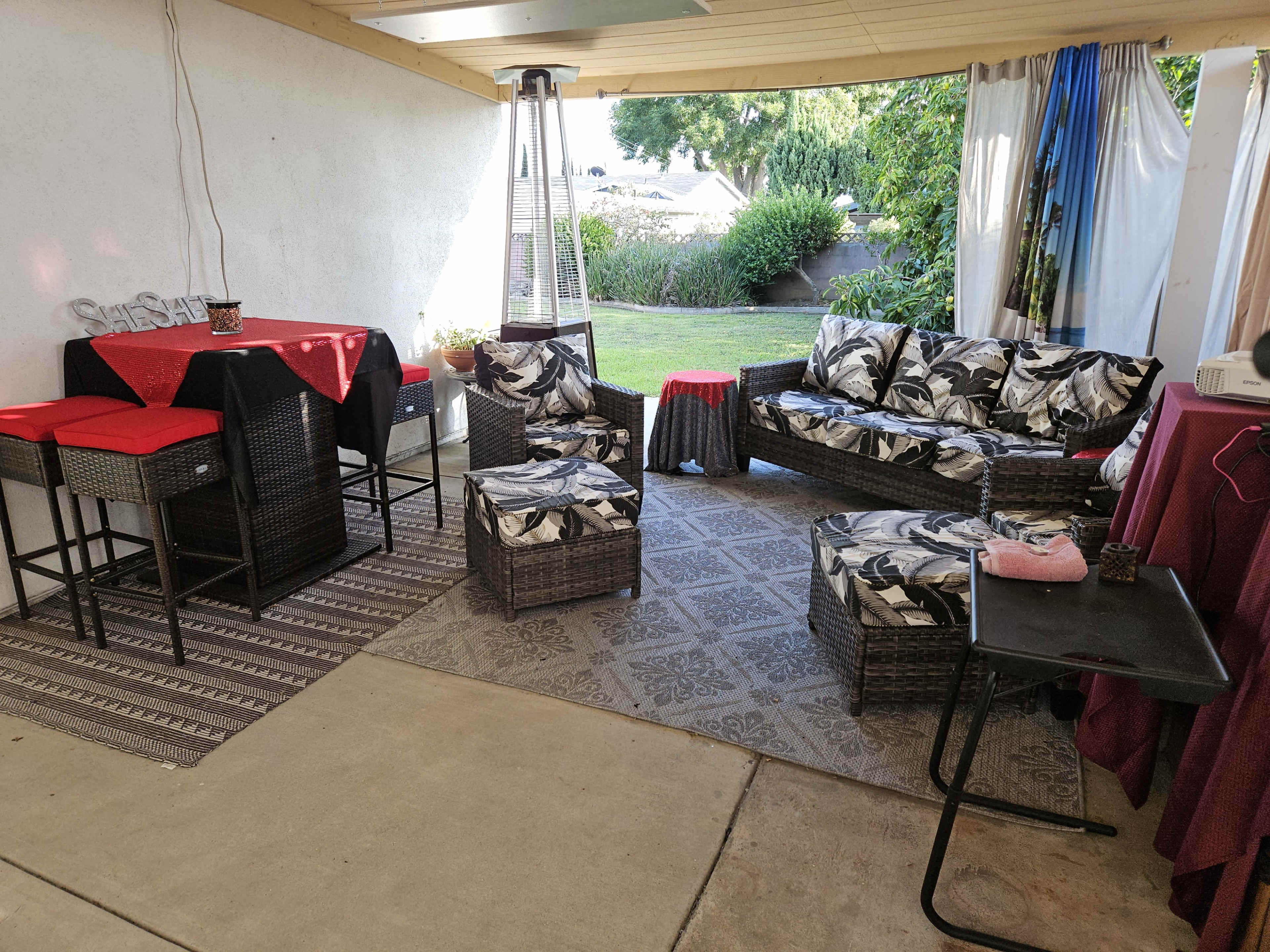A covered patio area featuring wicker furniture with floral cushions, a tall outdoor heater, and a small table, surrounded by greenery.