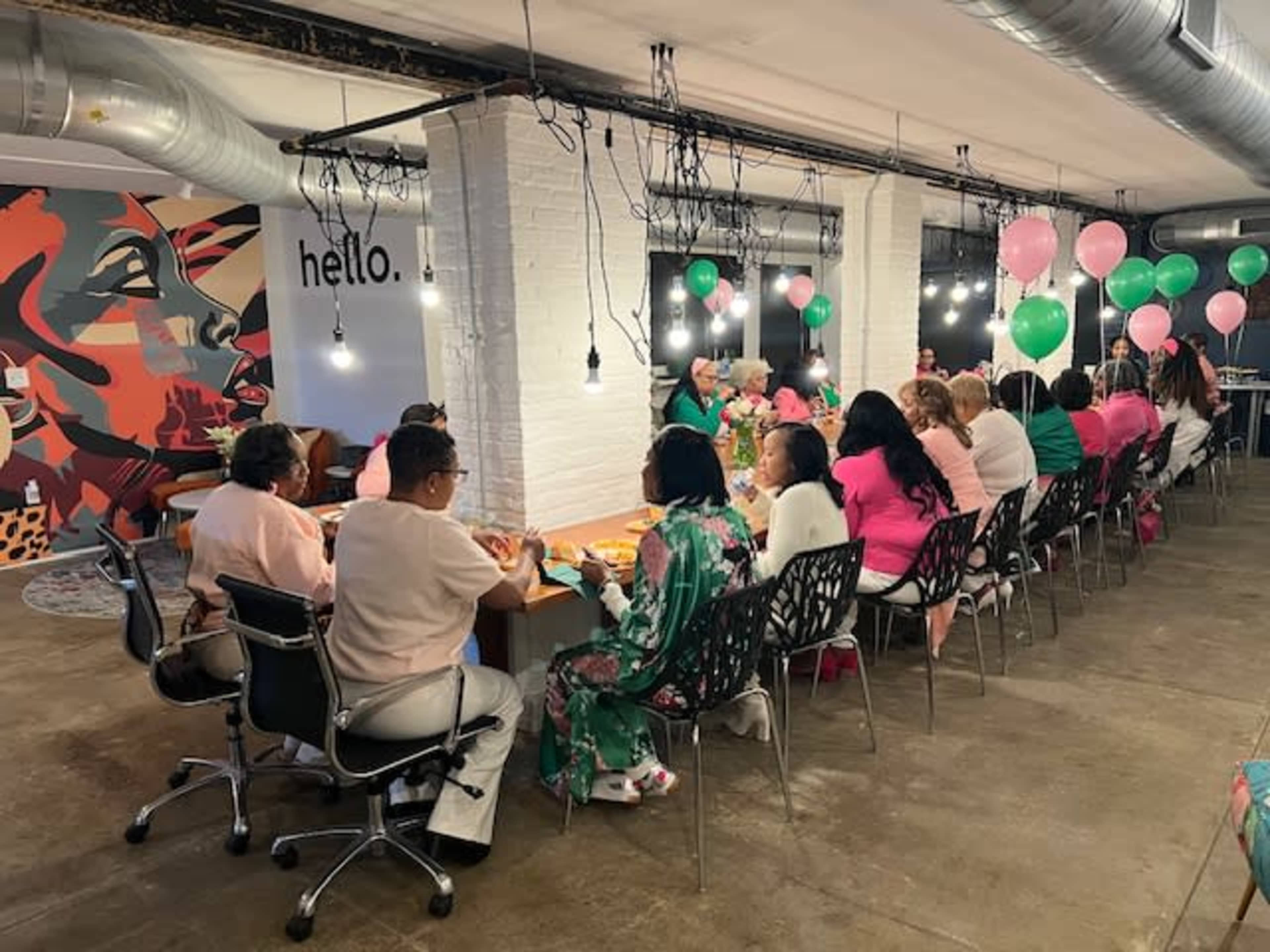 A large group of people is seated at a long table in a brightly lit room, decorated with pink and green balloons, while enjoying a gathering surrounded by colorful wall art.