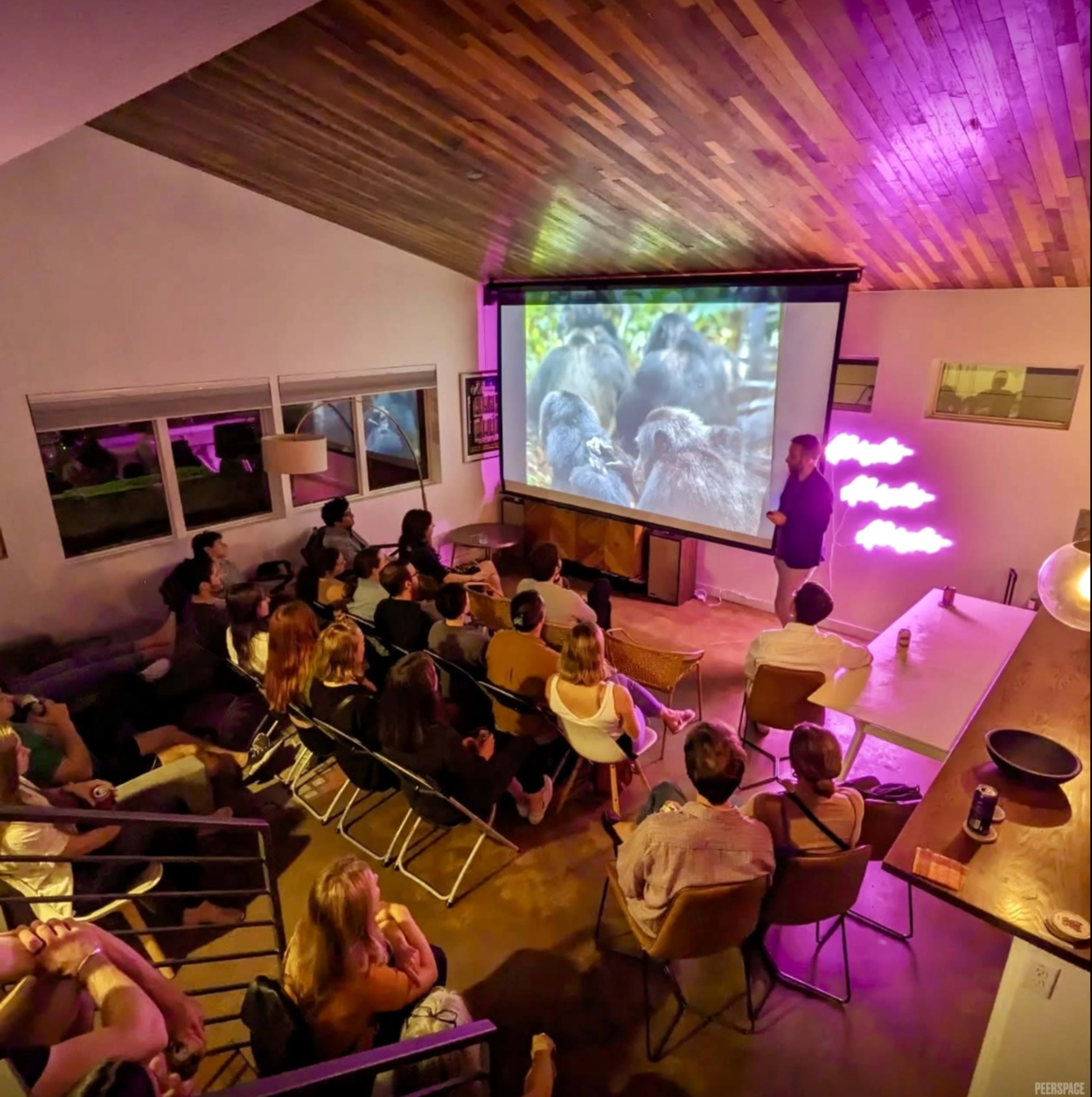 A speaker presents in front of a large screen displaying a wildlife image to an audience seated in a modern interior space.