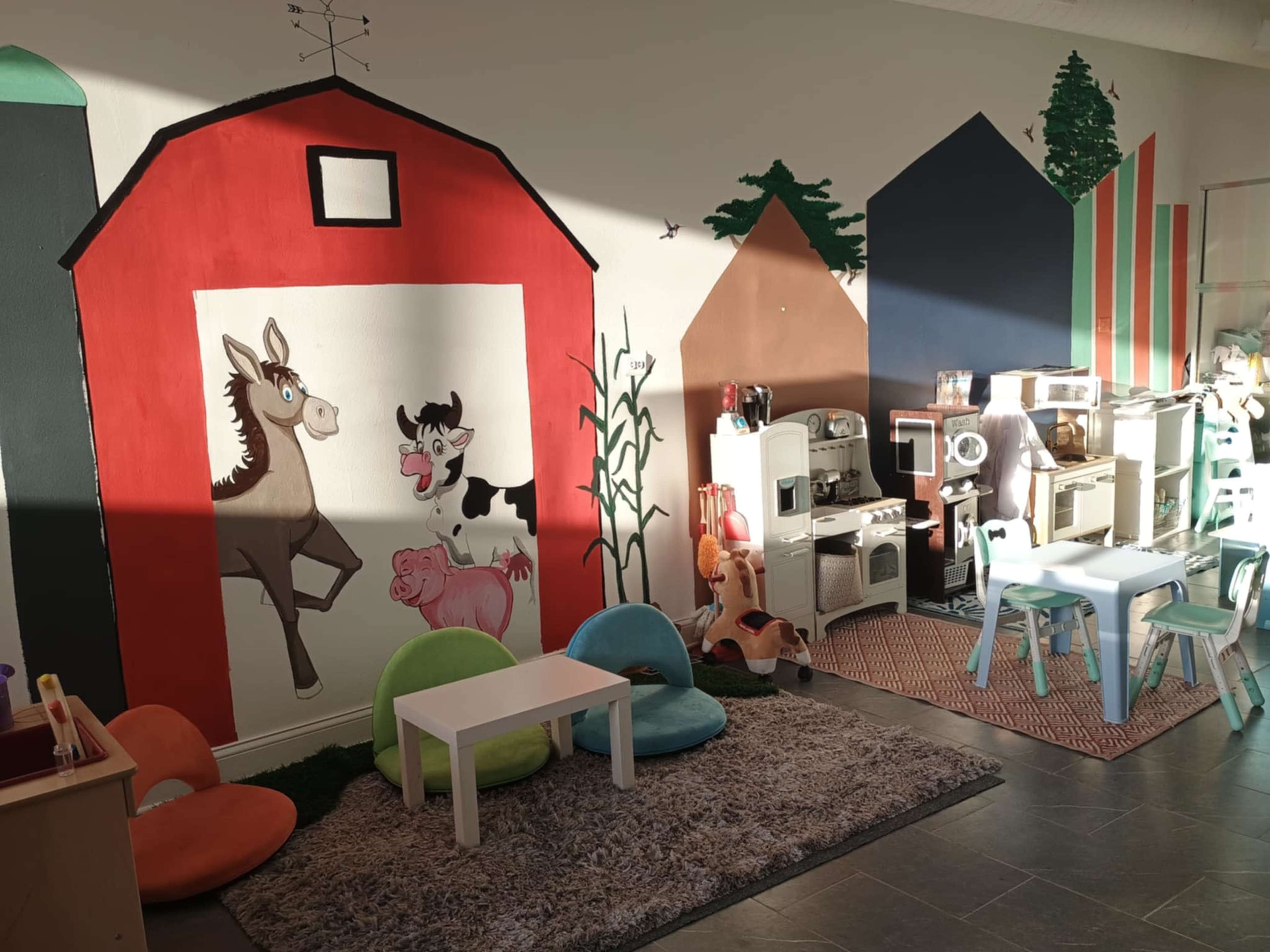 The image shows a brightly decorated children's playroom featuring murals of farm animals on one wall and various toy kitchen items arranged along the opposite wall.