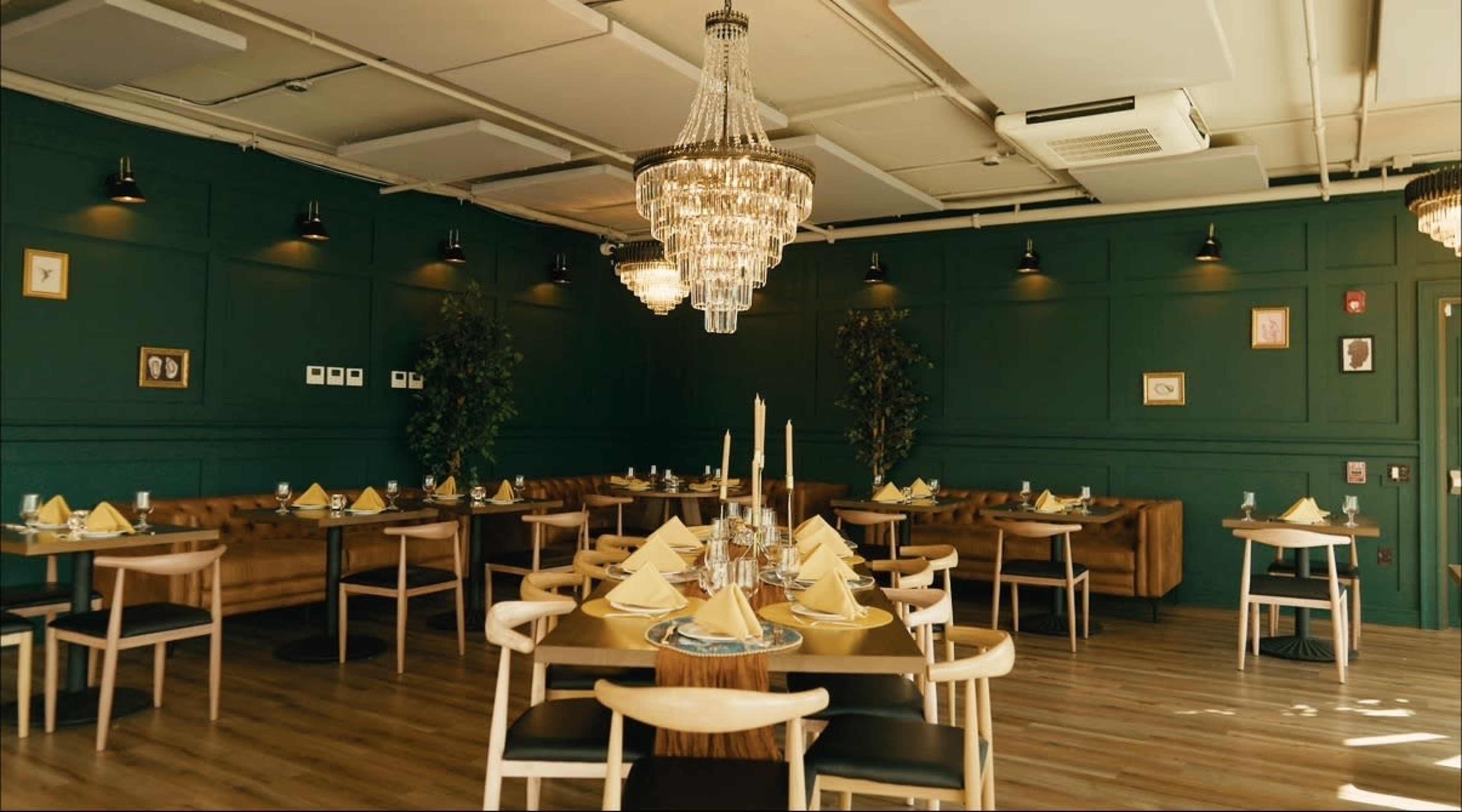 The image shows a well-decorated restaurant interior with dark green walls, elegant lighting, and tables set with yellow napkins and candles.