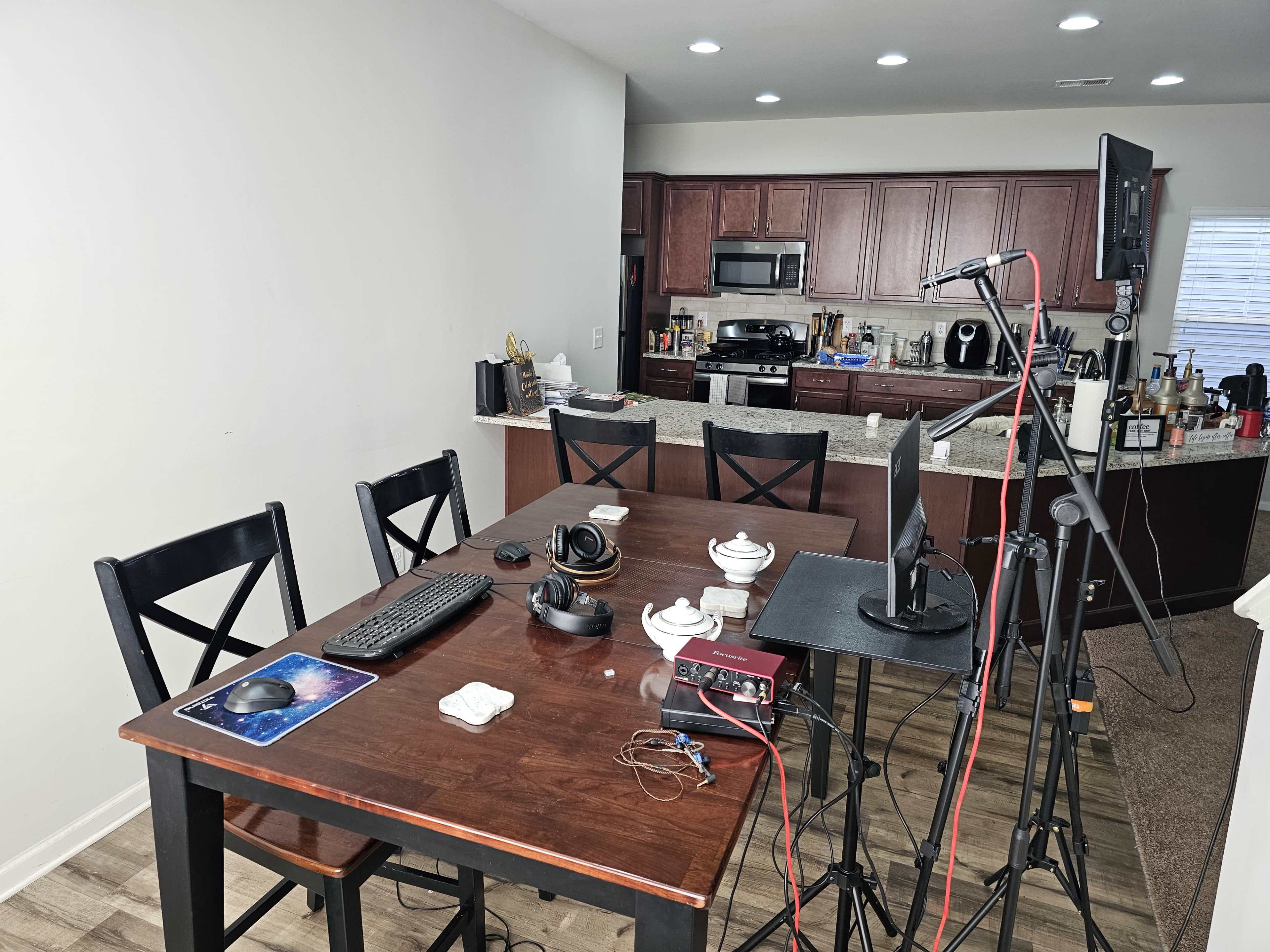 A dining table with a computer setup and various equipment is positioned near a kitchen with wooden cabinets and countertops.