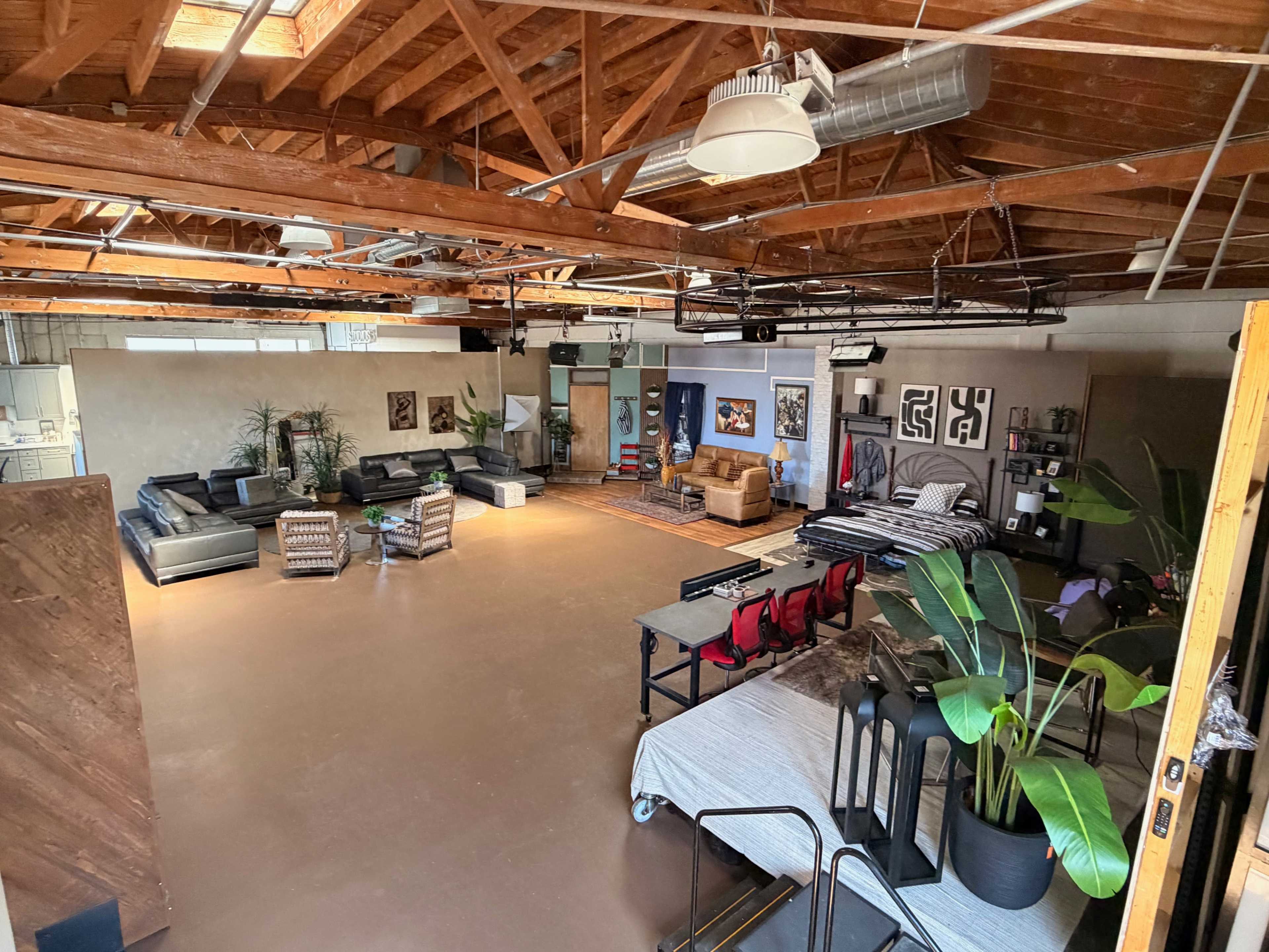 The image shows a spacious, open-layout studio with wooden beams, various seating arrangements, and decorative plants scattered throughout.