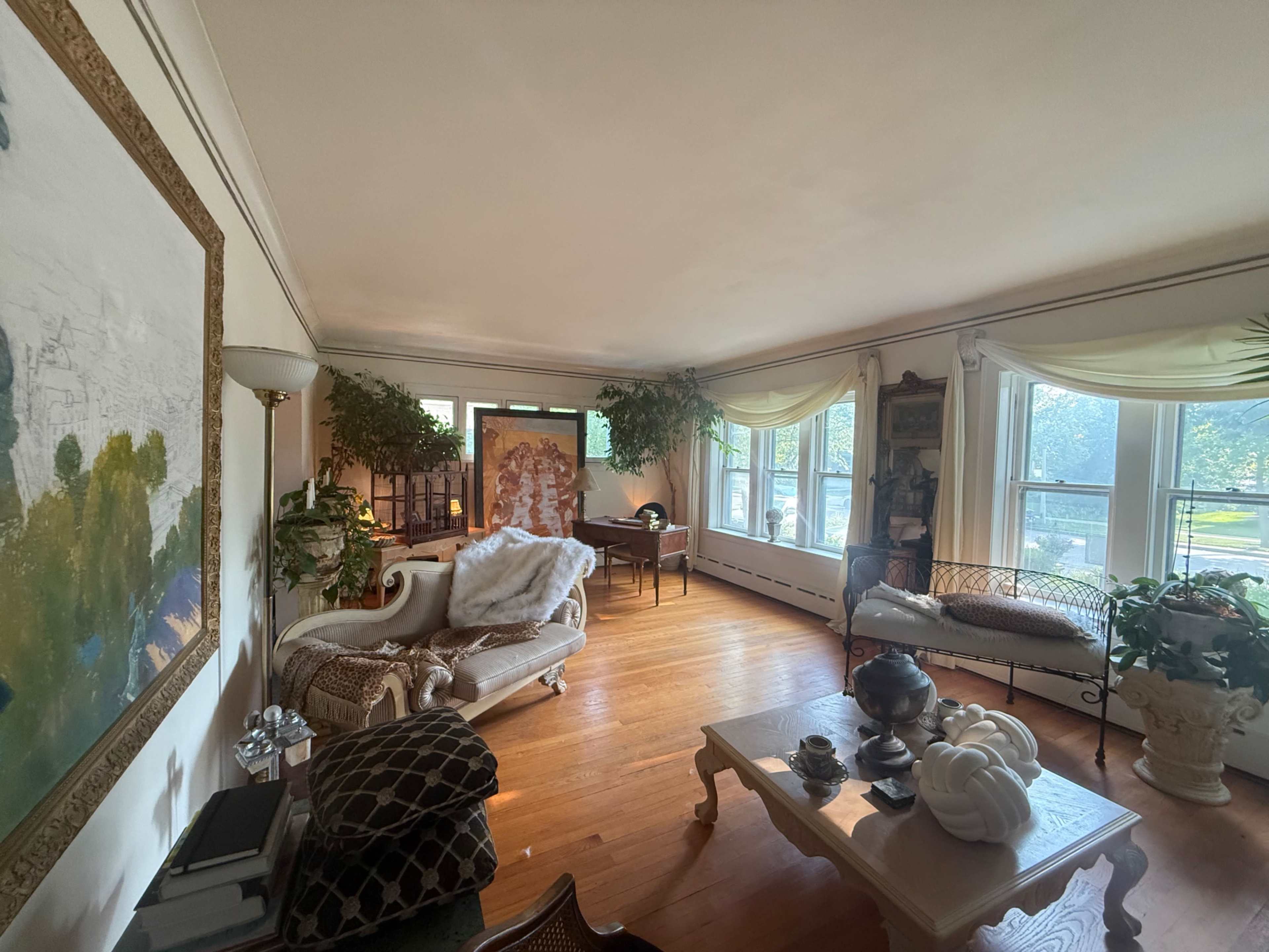 A spacious living room features a mix of furniture, including a sofa, a wrought-iron bench, and a wooden table, surrounded by large windows and various decorative plants.