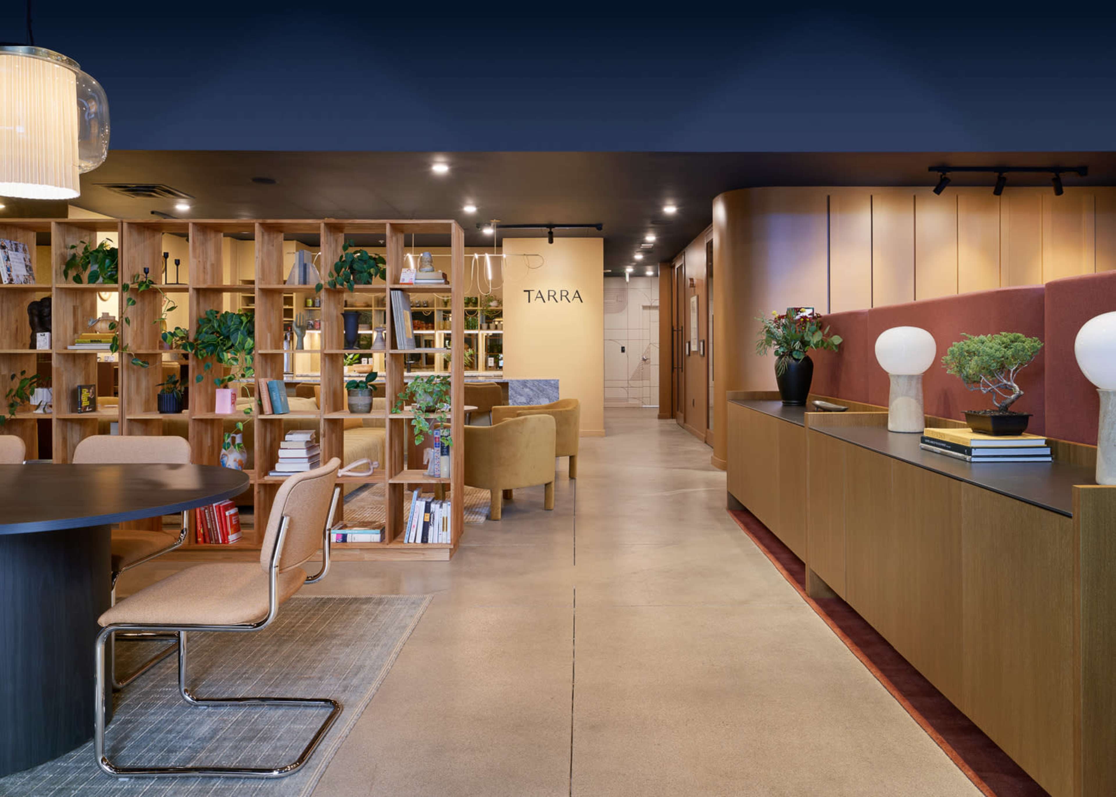 A modern interior space featuring wooden bookshelves filled with books and plants, a round table, and a long counter with decorative items and seating areas.