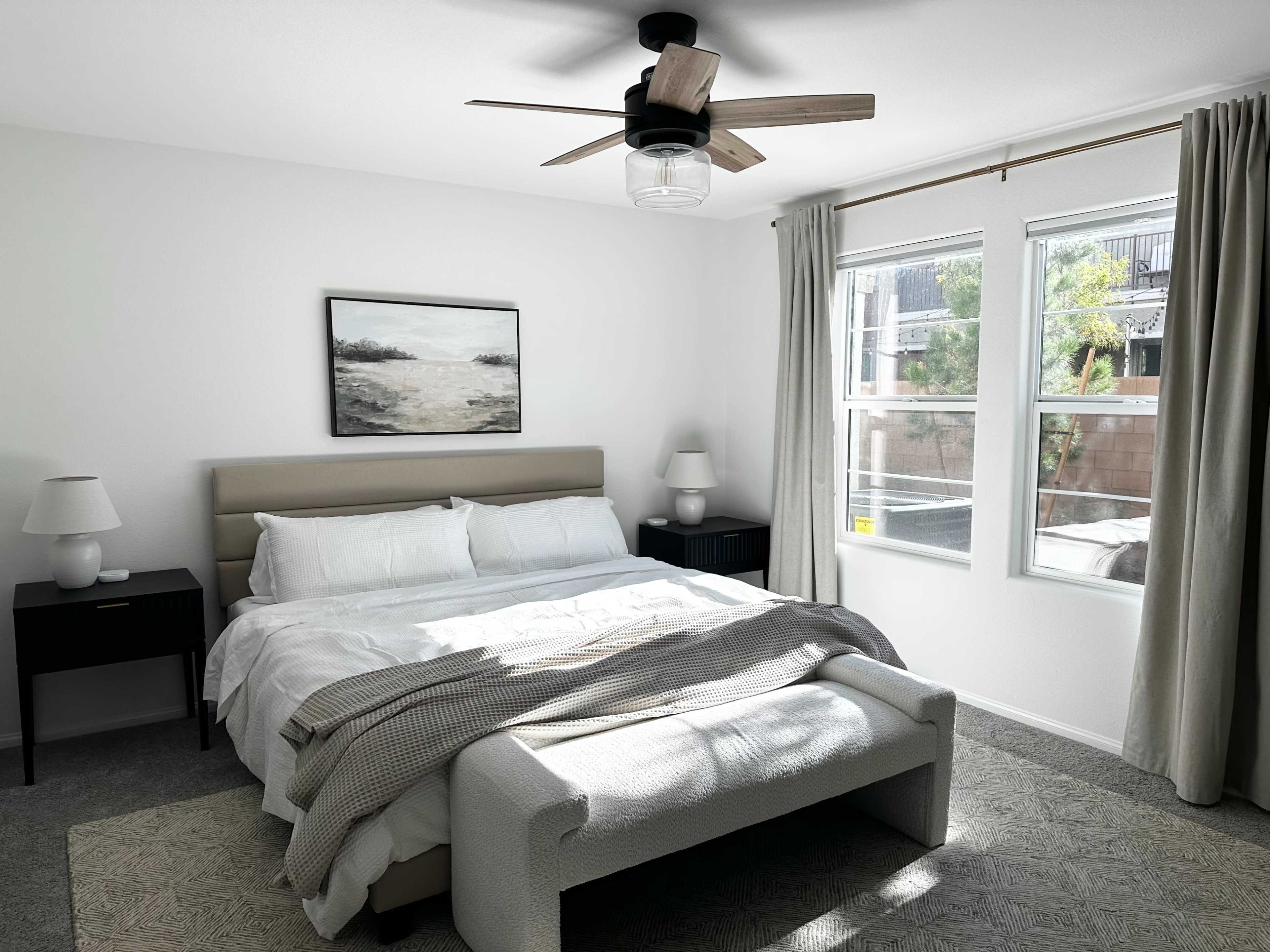 The image shows a neatly arranged bedroom featuring a bed with a light-colored duvet, two nightstands, a ceiling fan, and large windows letting in natural light.