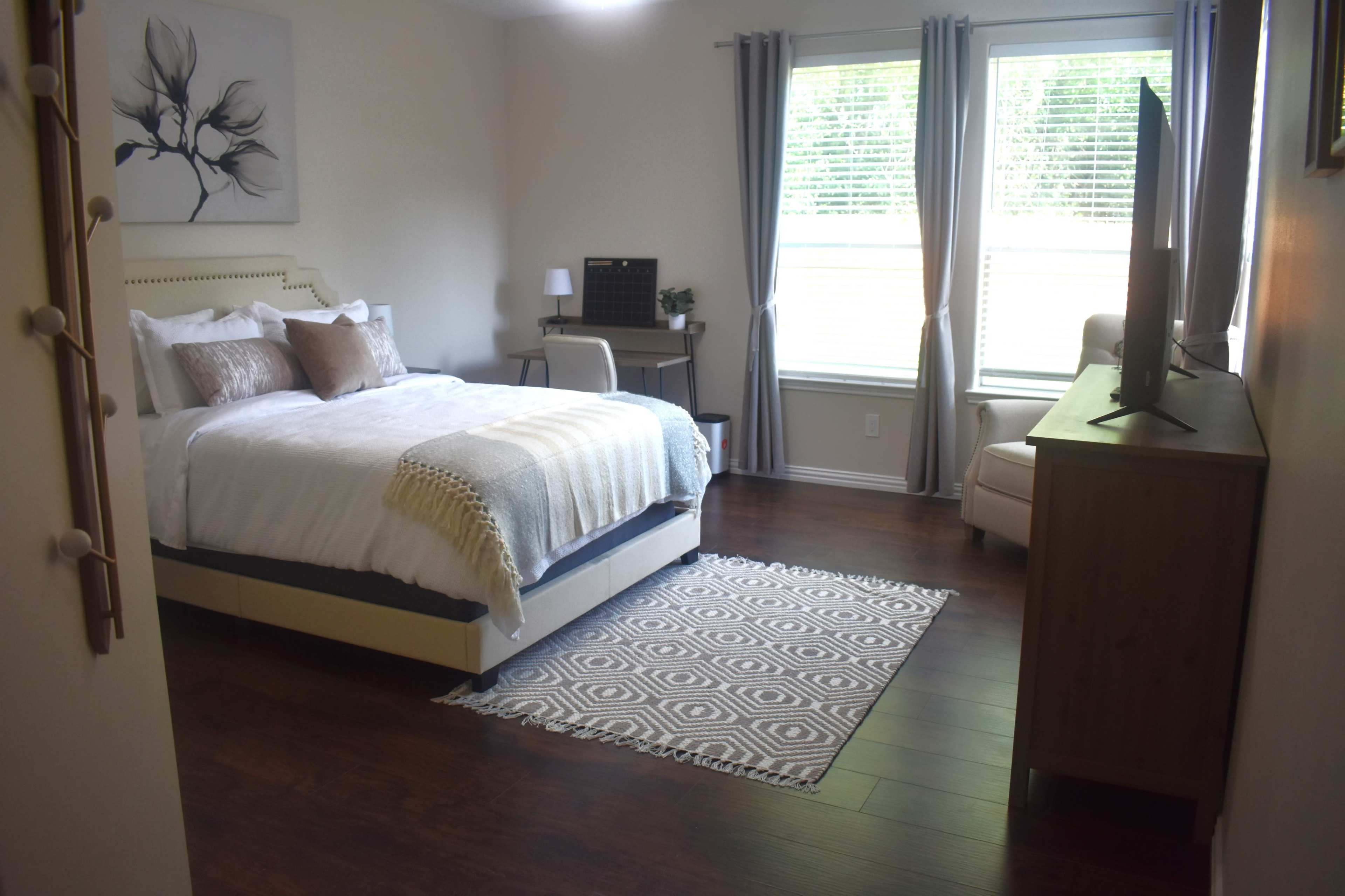 A neatly arranged bedroom featuring a bed, a desk, a chair, and a television, with natural light streaming through two windows.
