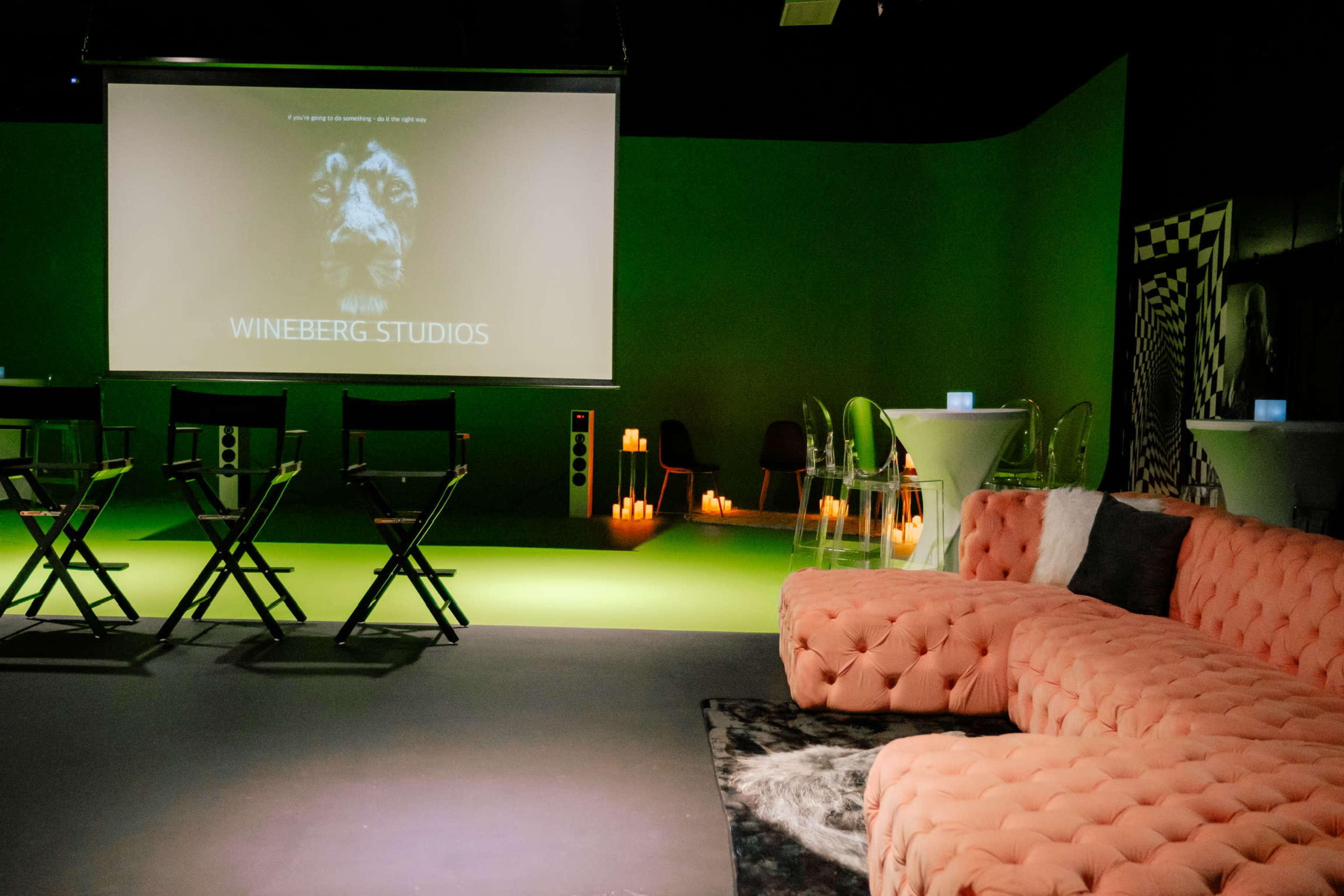 The image shows a modern studio space with a green accent wall, a large screen displaying "WINEBERG STUDIOS," and furniture including a pink sectional sofa and folding chairs arranged around tables with candles.