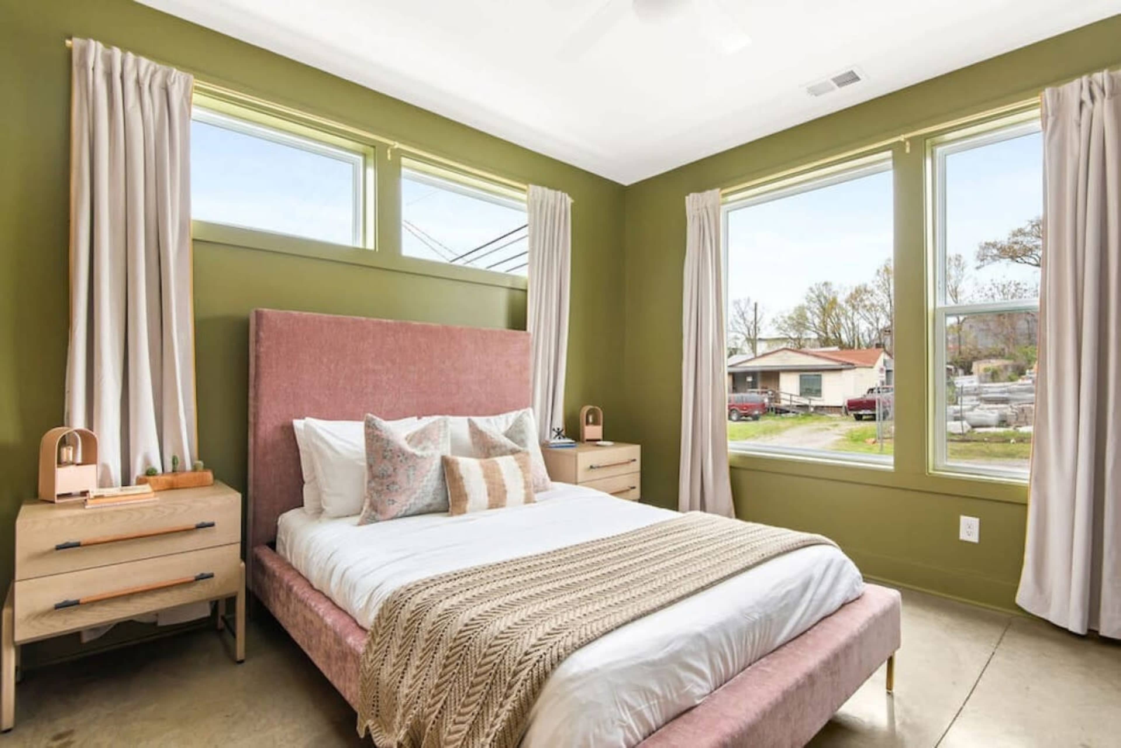 A bedroom with a pink upholstered bed, light-colored bedding, wooden nightstands, and large windows that provide natural light.