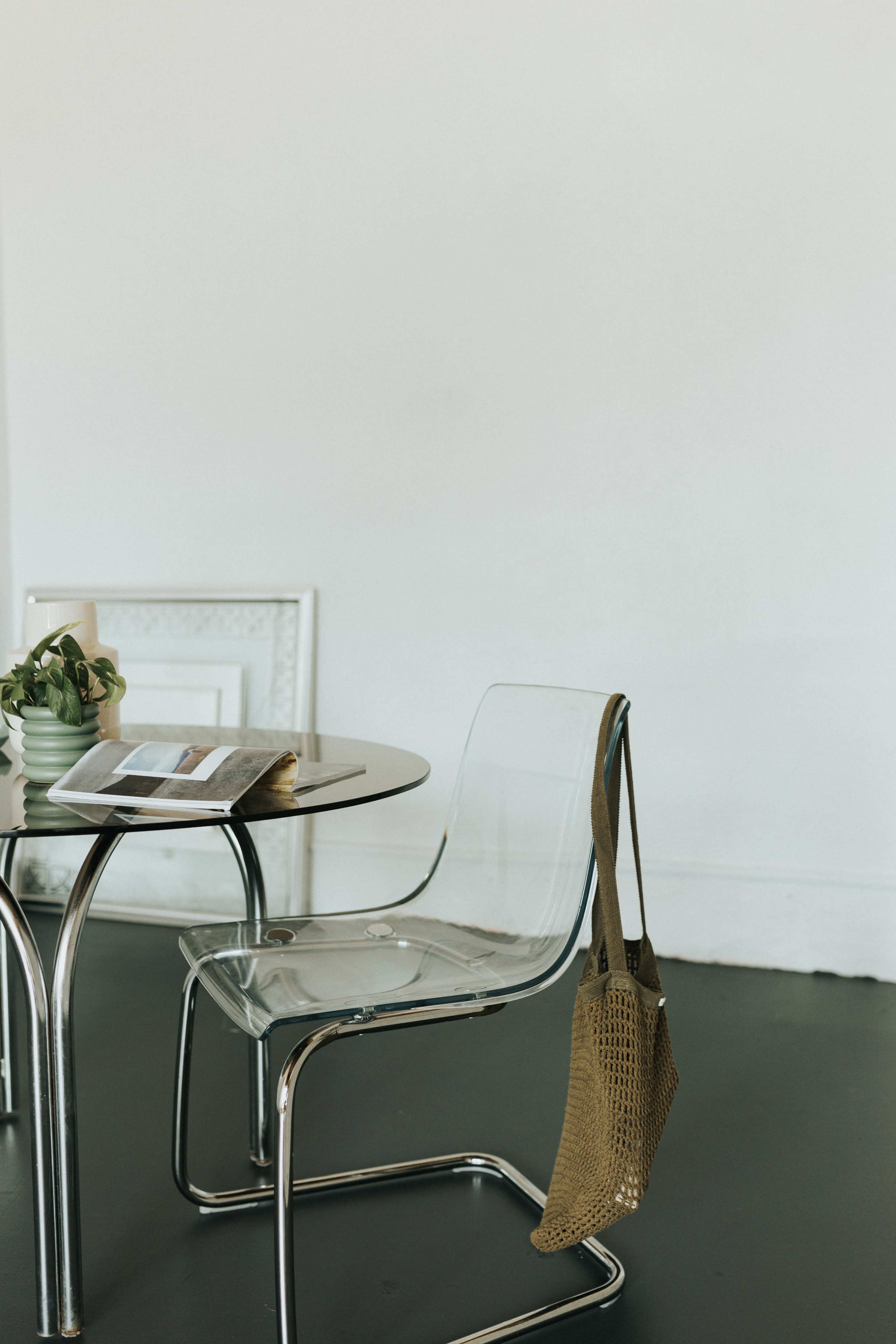 A transparent chair with a tan tote bag hangs on the side next to a round glass table and a small potted plant.