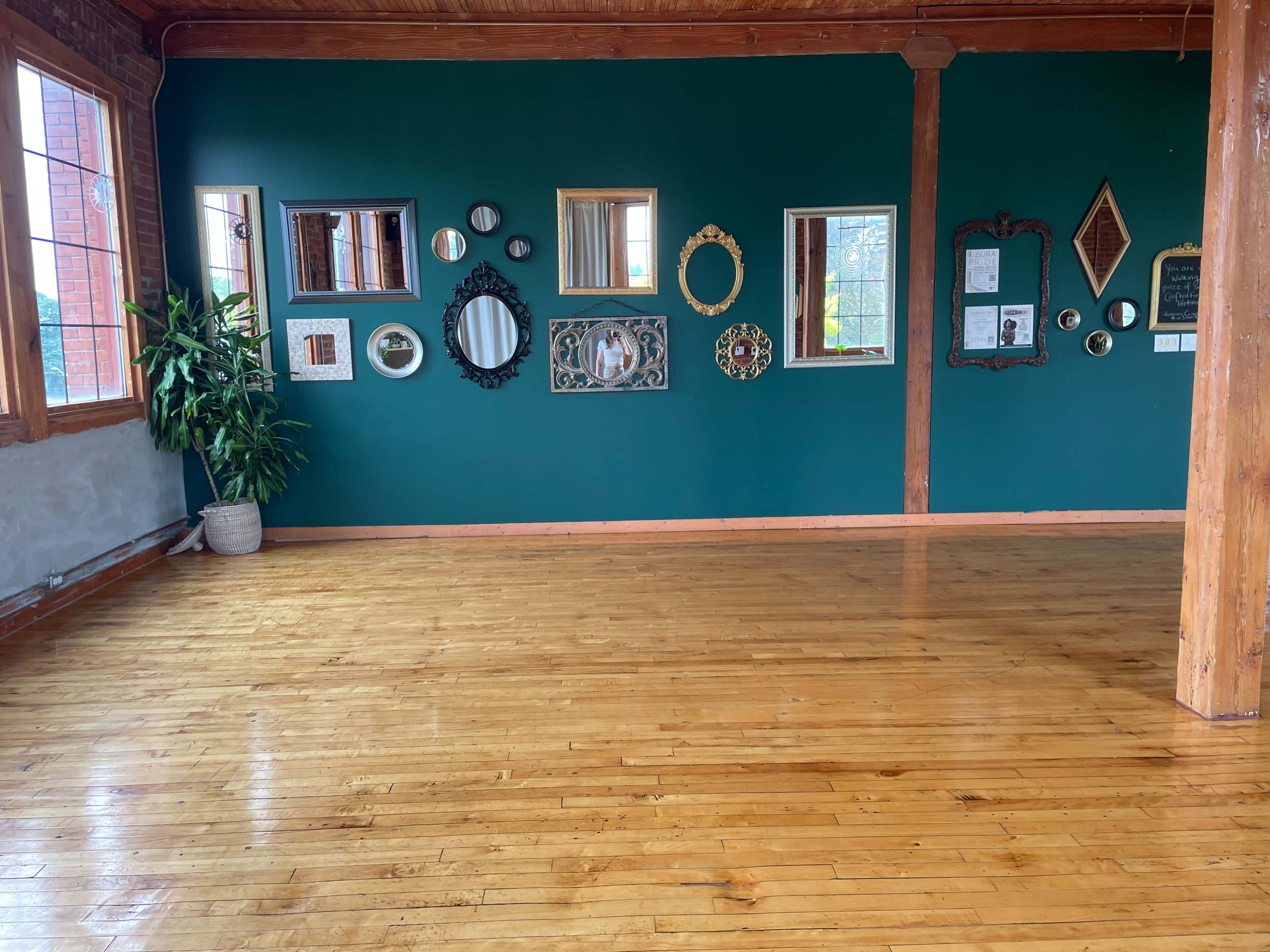 The image shows a spacious room with a blue-green wall adorned with various decorative mirrors, wooden flooring, and a potted plant in the corner.
