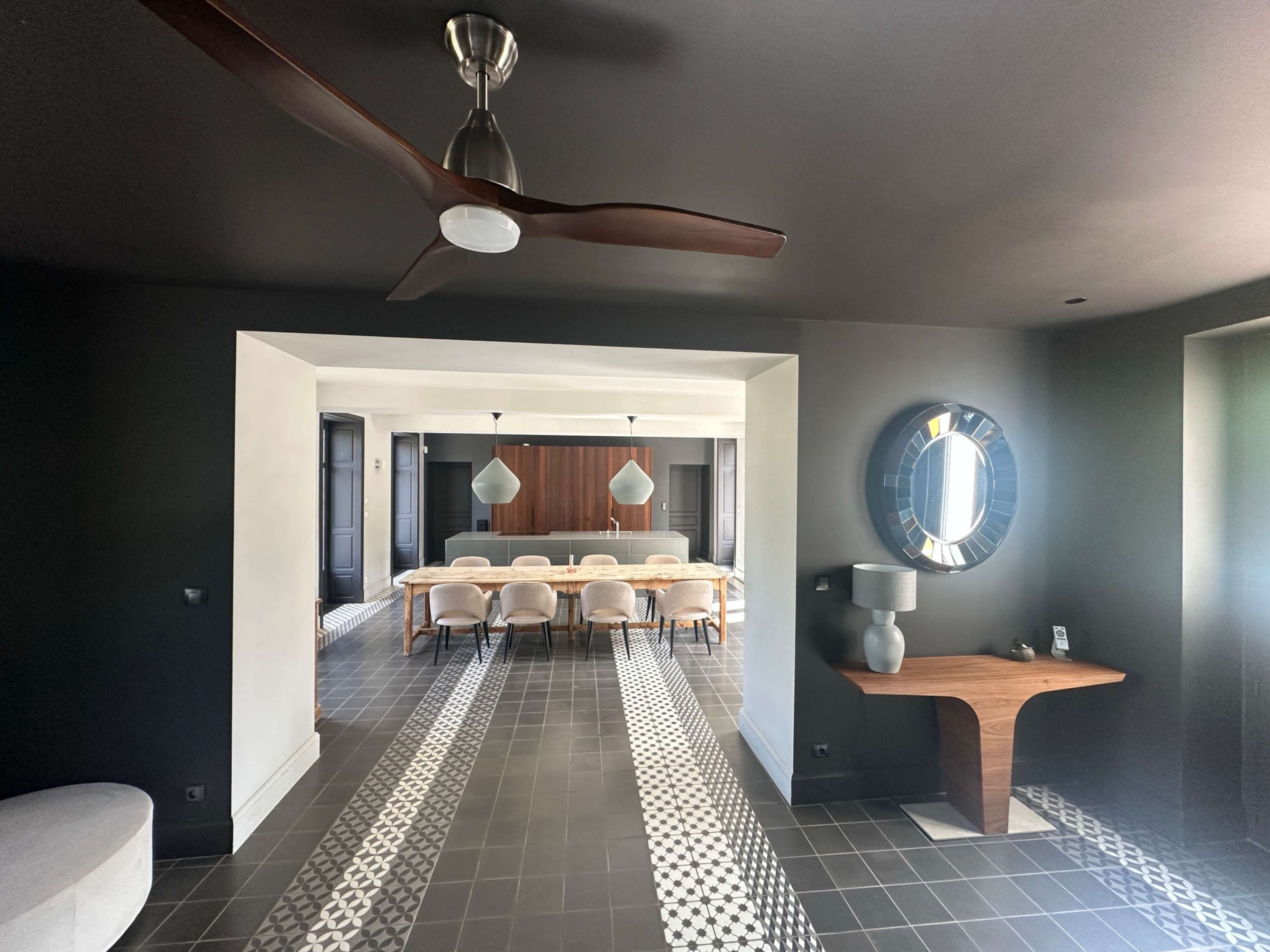 The image shows an interior space featuring a modern dining area with a long table, surrounded by chairs, and a sleek design that includes dark walls and decorative flooring.