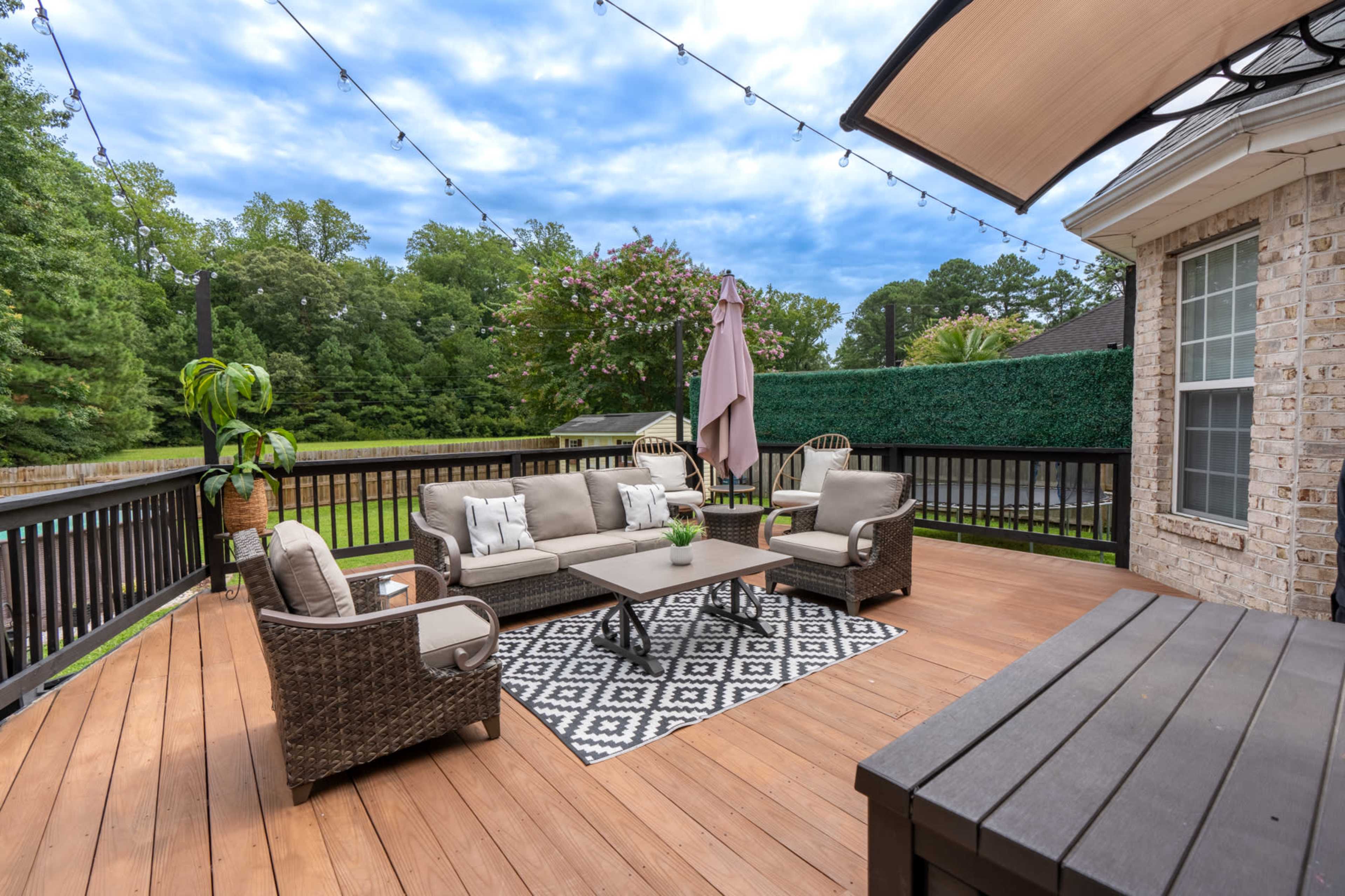 A wooden deck furnished with a seating area that includes a sofa, two armchairs, and a coffee table, surrounded by greenery and string lights overhead.
