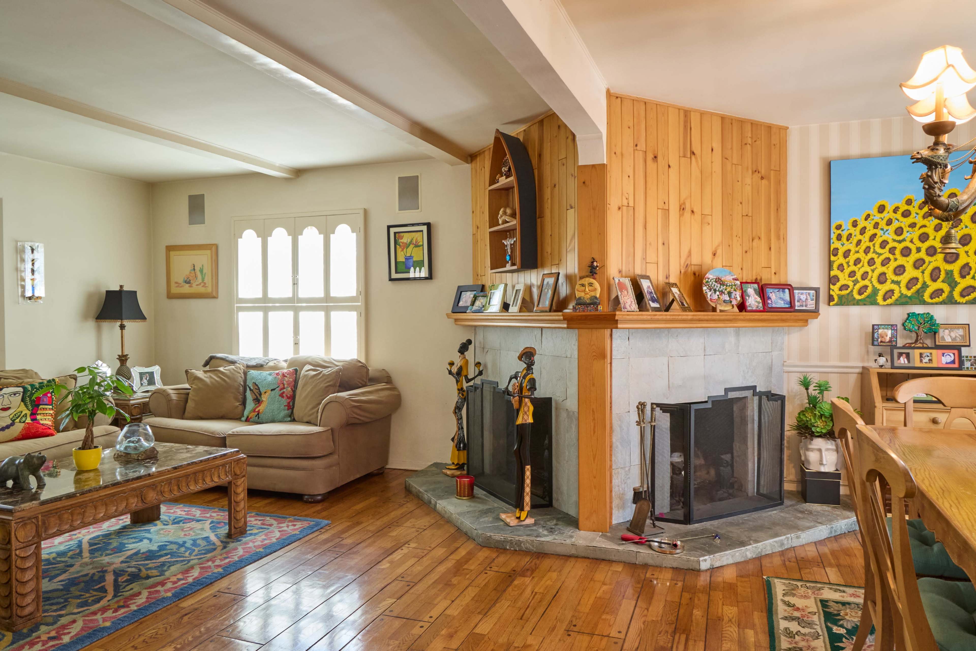 The image shows a cozy living room featuring a light-colored sofa, a wooden fireplace with decorative items, and a dining area with a colorful artwork on the wall.