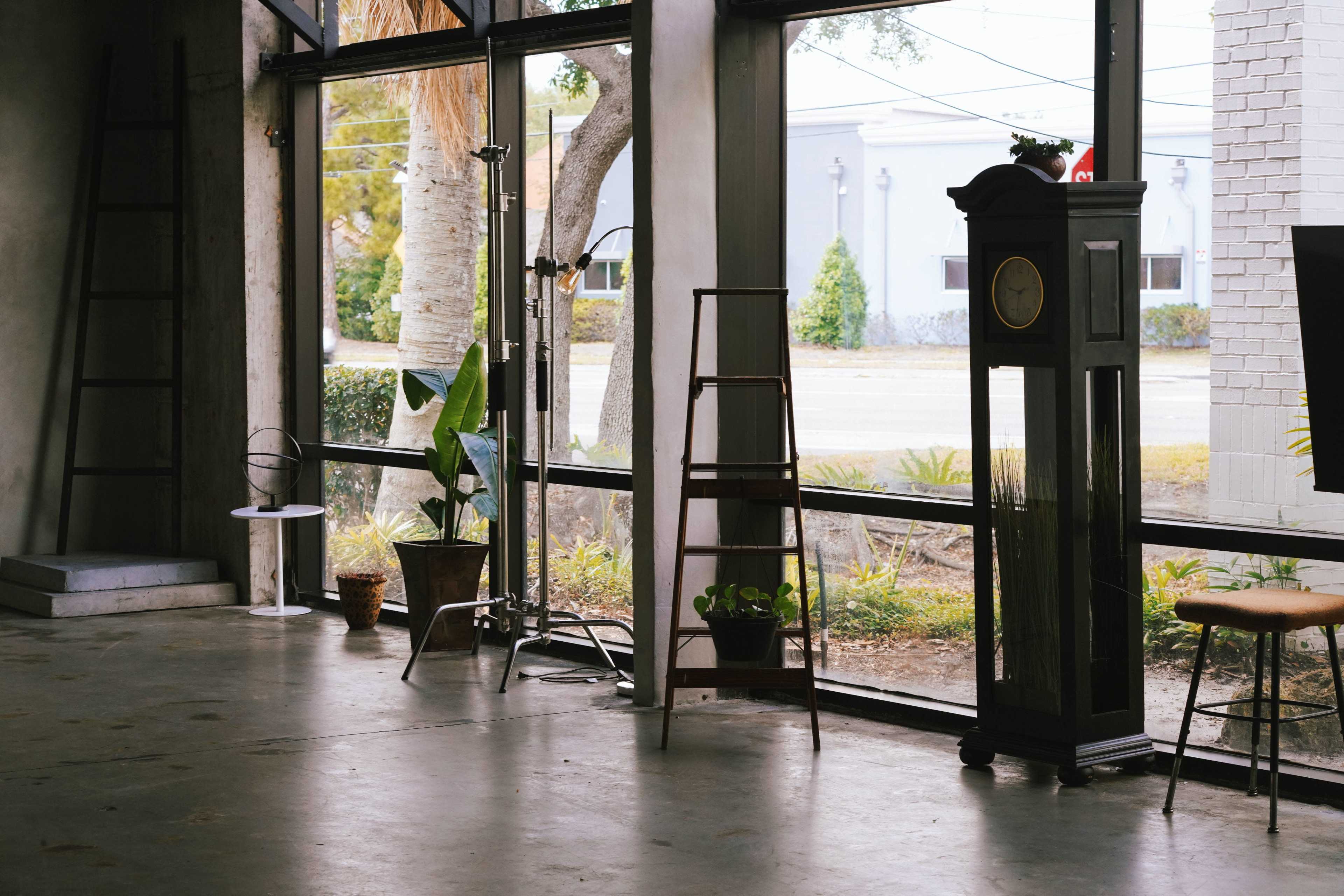 The image shows an empty interior space with large windows, a few pieces of furniture, including a clock and a ladder, and a view of the outdoors featuring trees and a building.