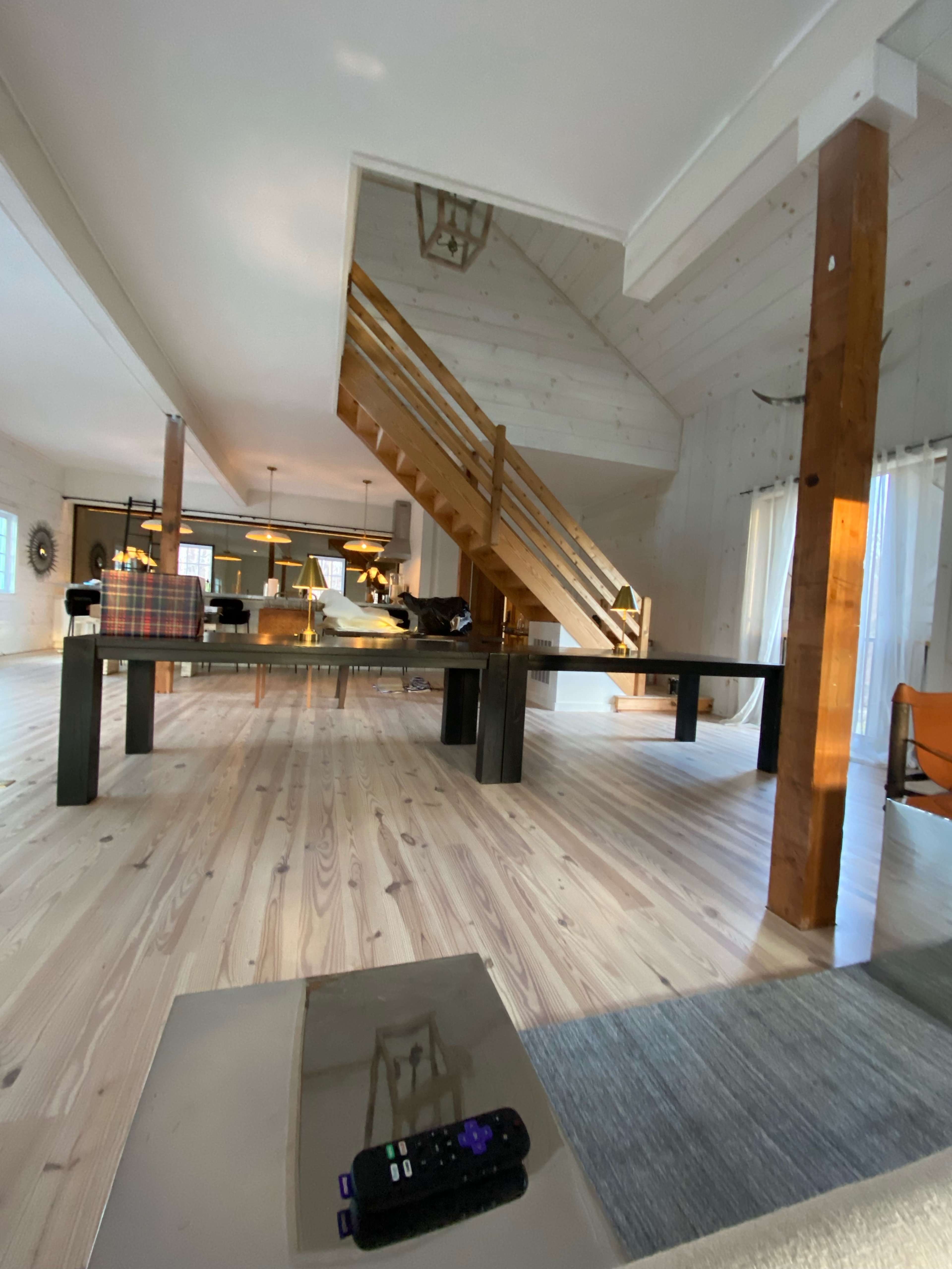 The image shows a spacious interior with wooden flooring, a staircase, and a large dining table set in a well-lit area.