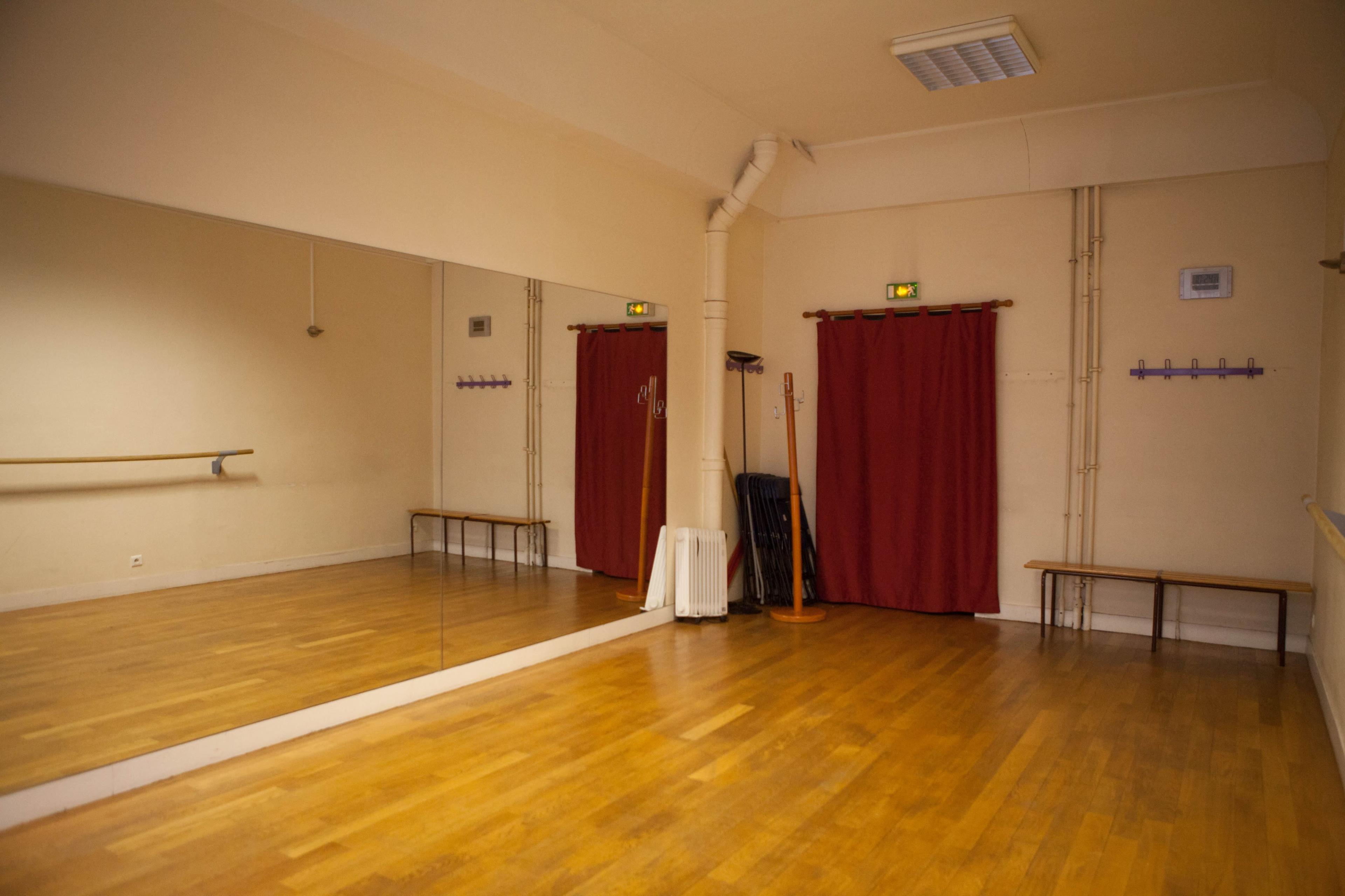 The image depicts a spacious dance studio featuring a large mirror, red curtains, wooden flooring, and minimalist furnishings.