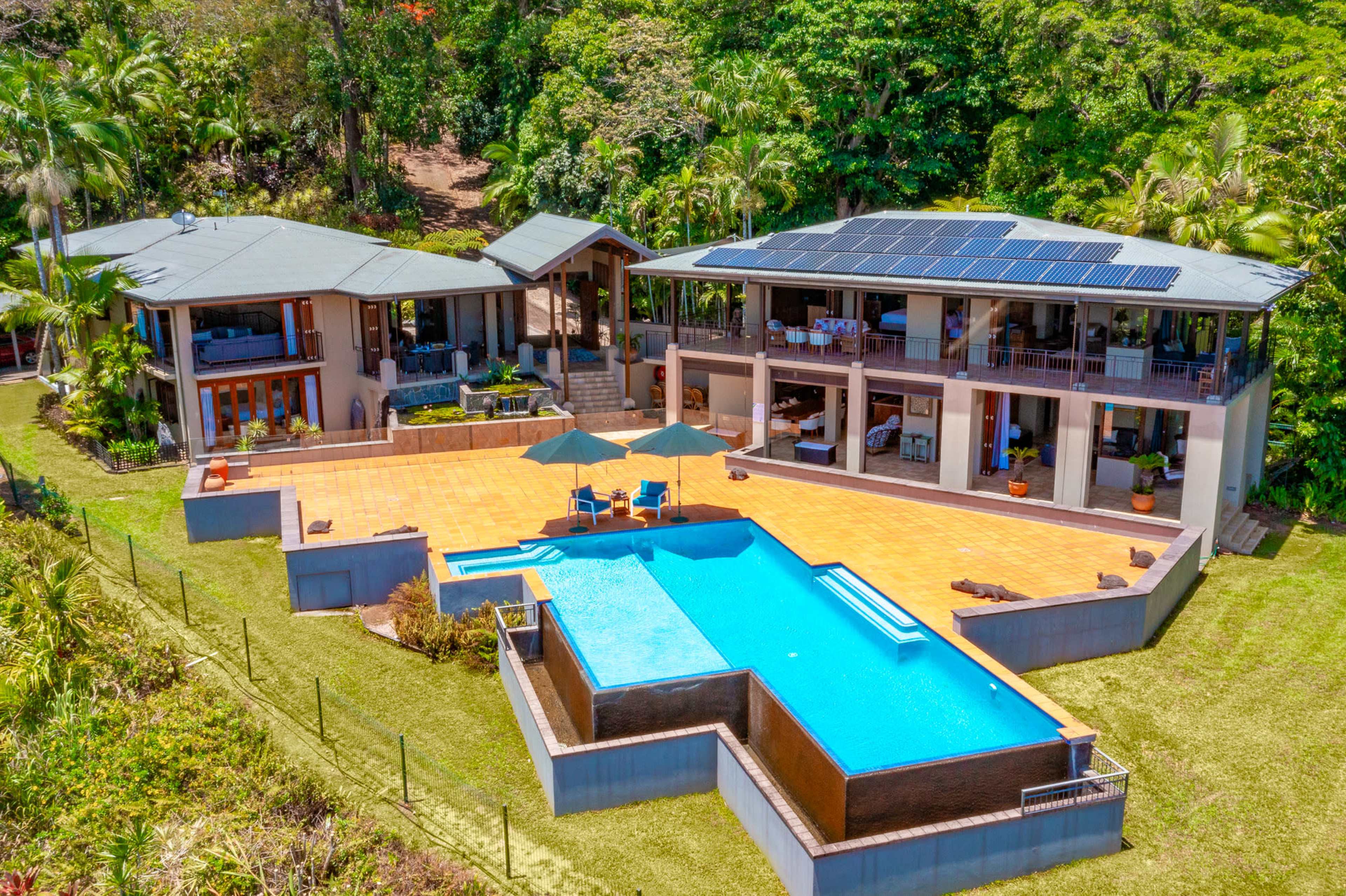 The image shows a modern two-story house with solar panels, surrounded by lush greenery and featuring a large swimming pool and a patio area.