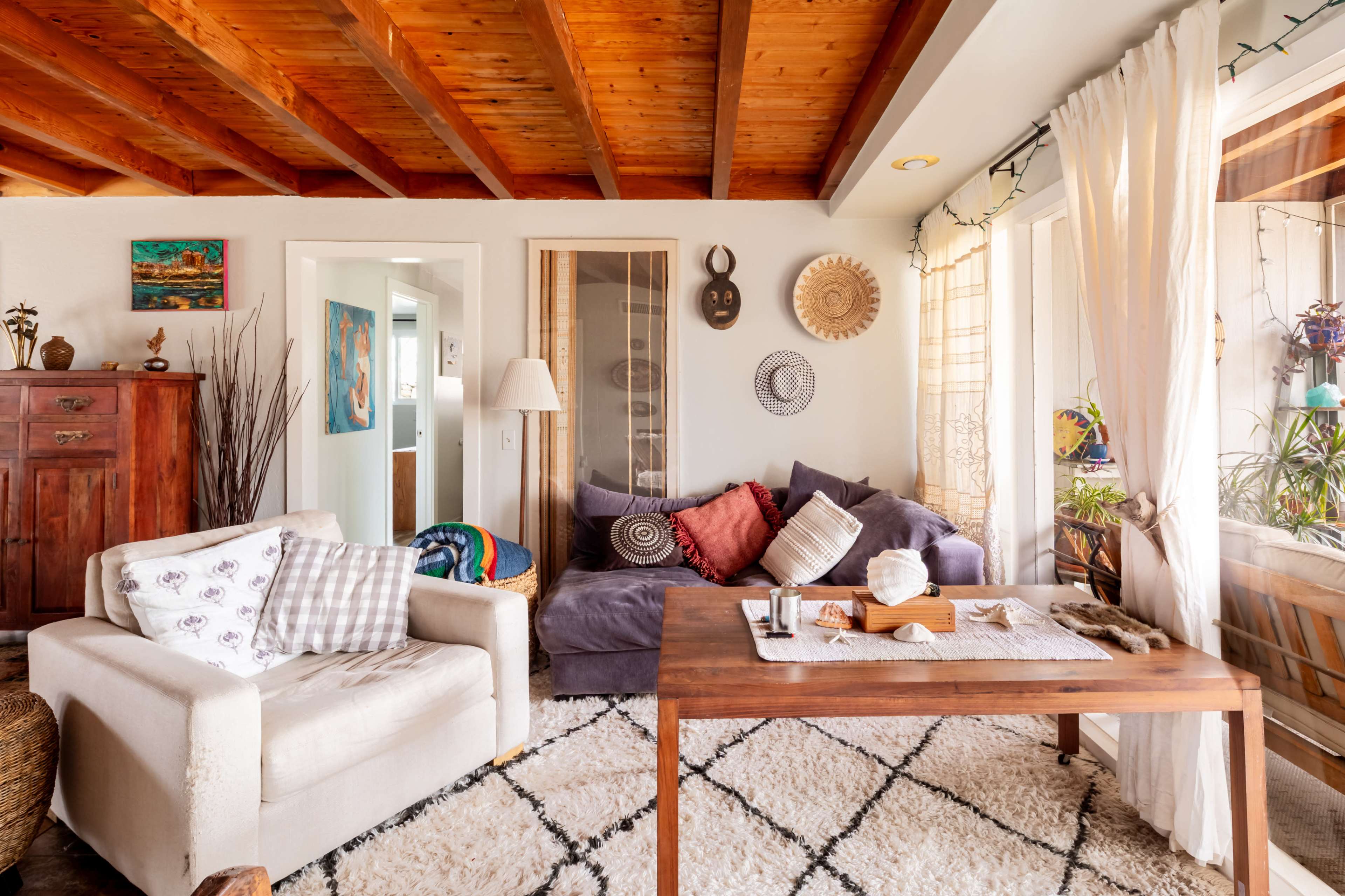 The image shows a cozy living room with a wooden ceiling, a mix of comfortable seating, and decor that includes artwork and plants.