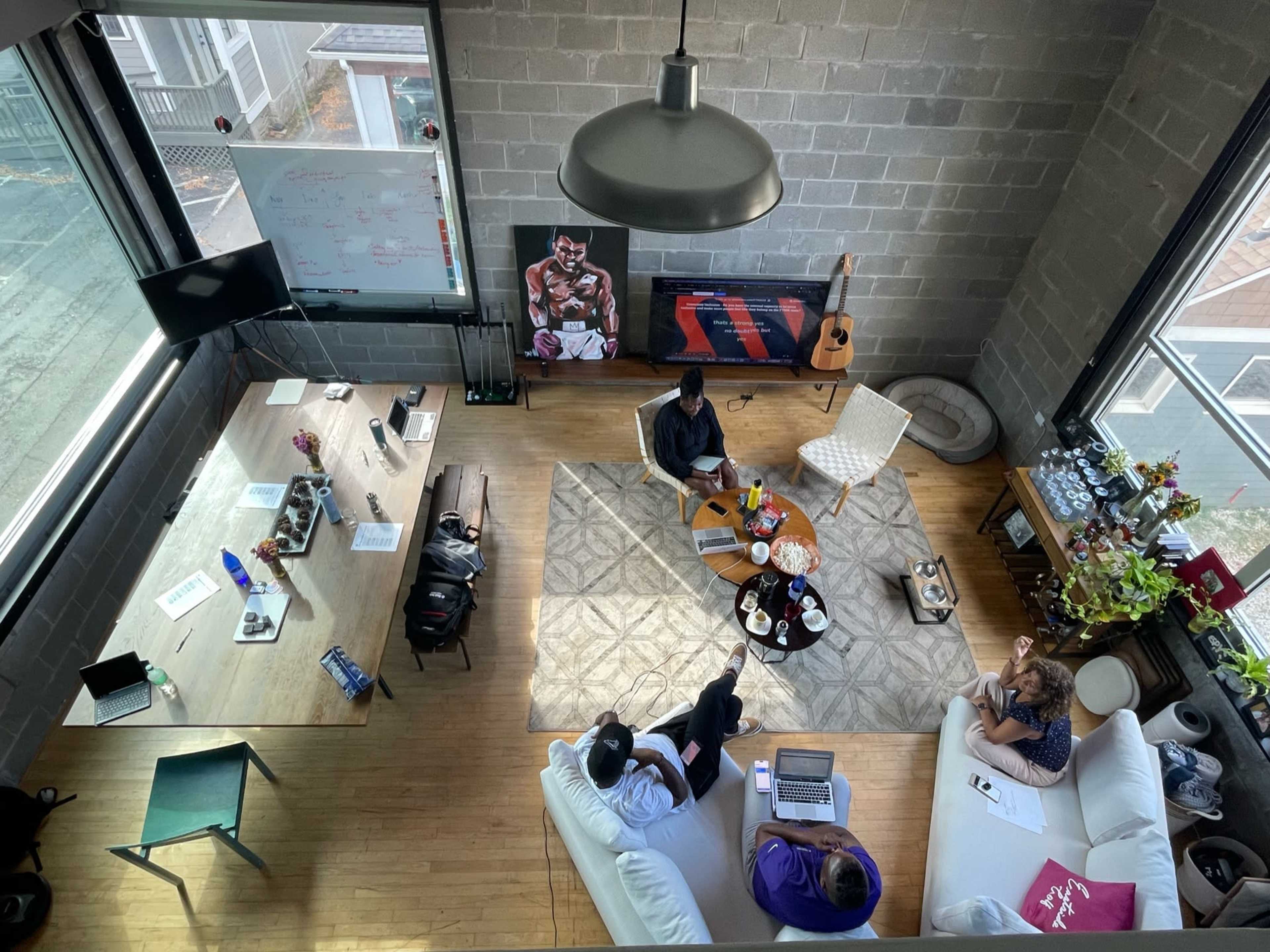 A modern workspace featuring a central seating area with three people discussing, surrounded by large windows and various work materials.