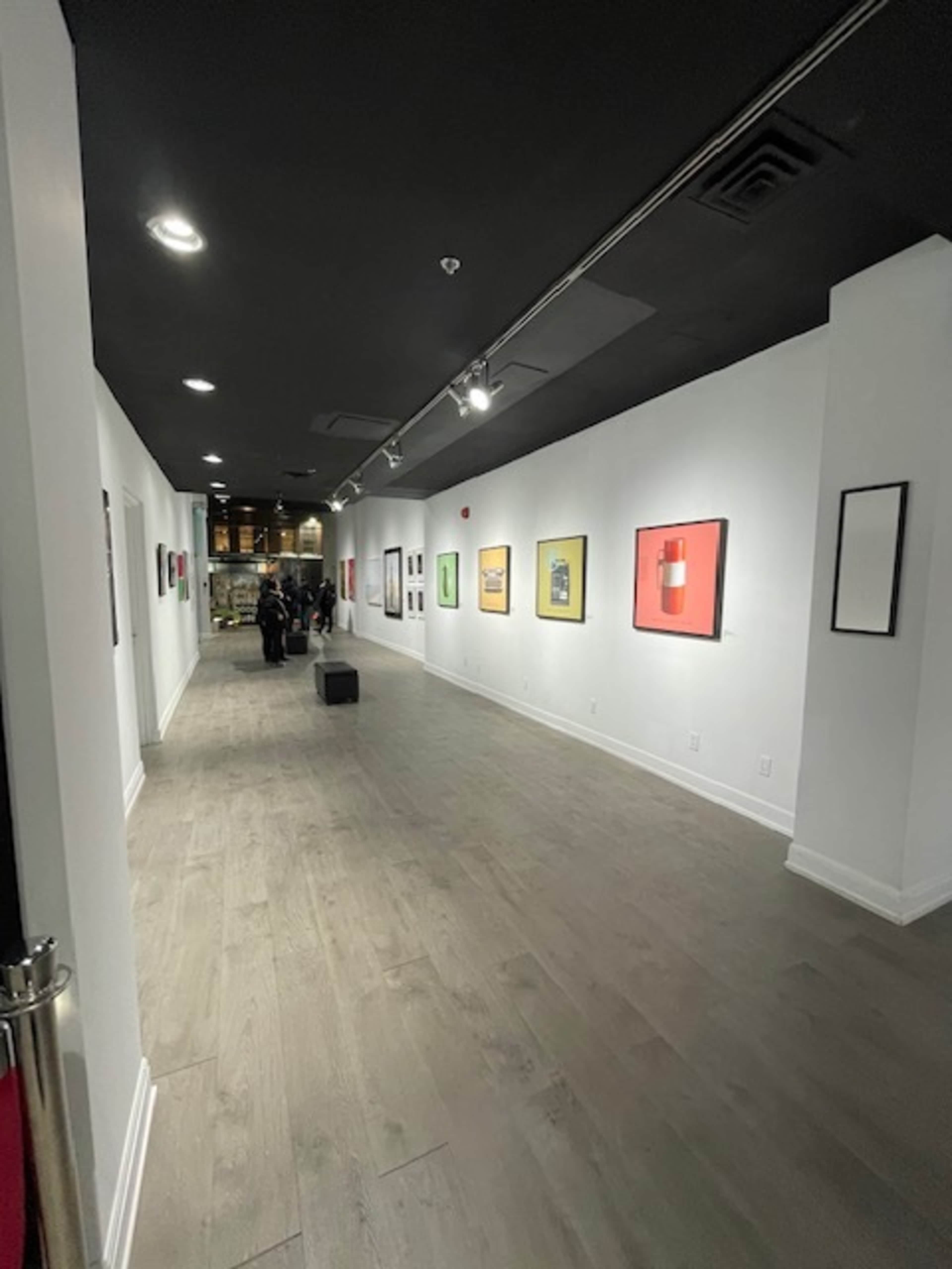 A long, well-lit art gallery hallway with framed artworks displayed on the walls and a group of people in the background.