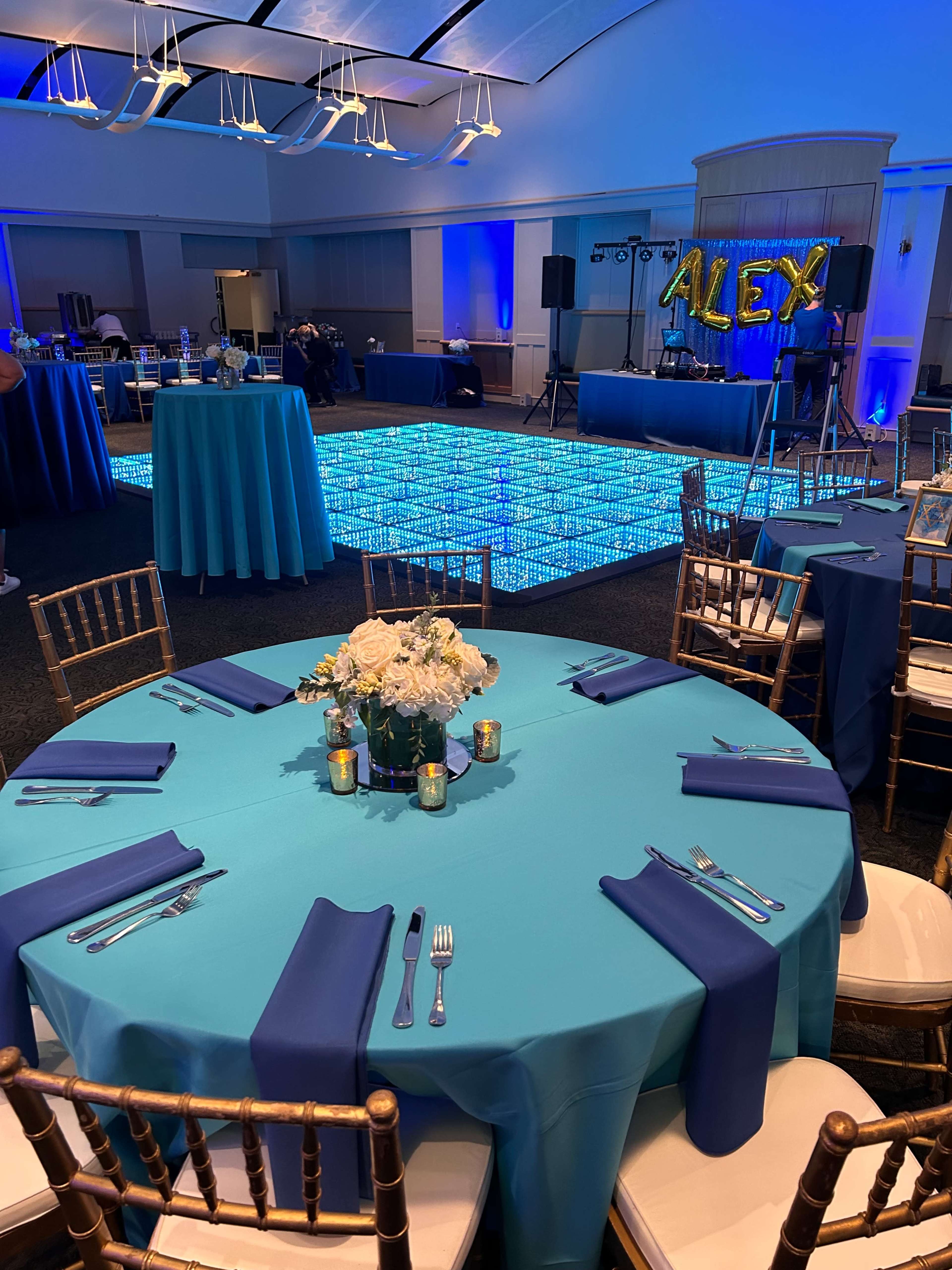 A decorated event space featuring round tables with turquoise tablecloths, blue napkins, and floral centerpieces, alongside a light-up dance floor and a stage with a "ALEX" balloon display.