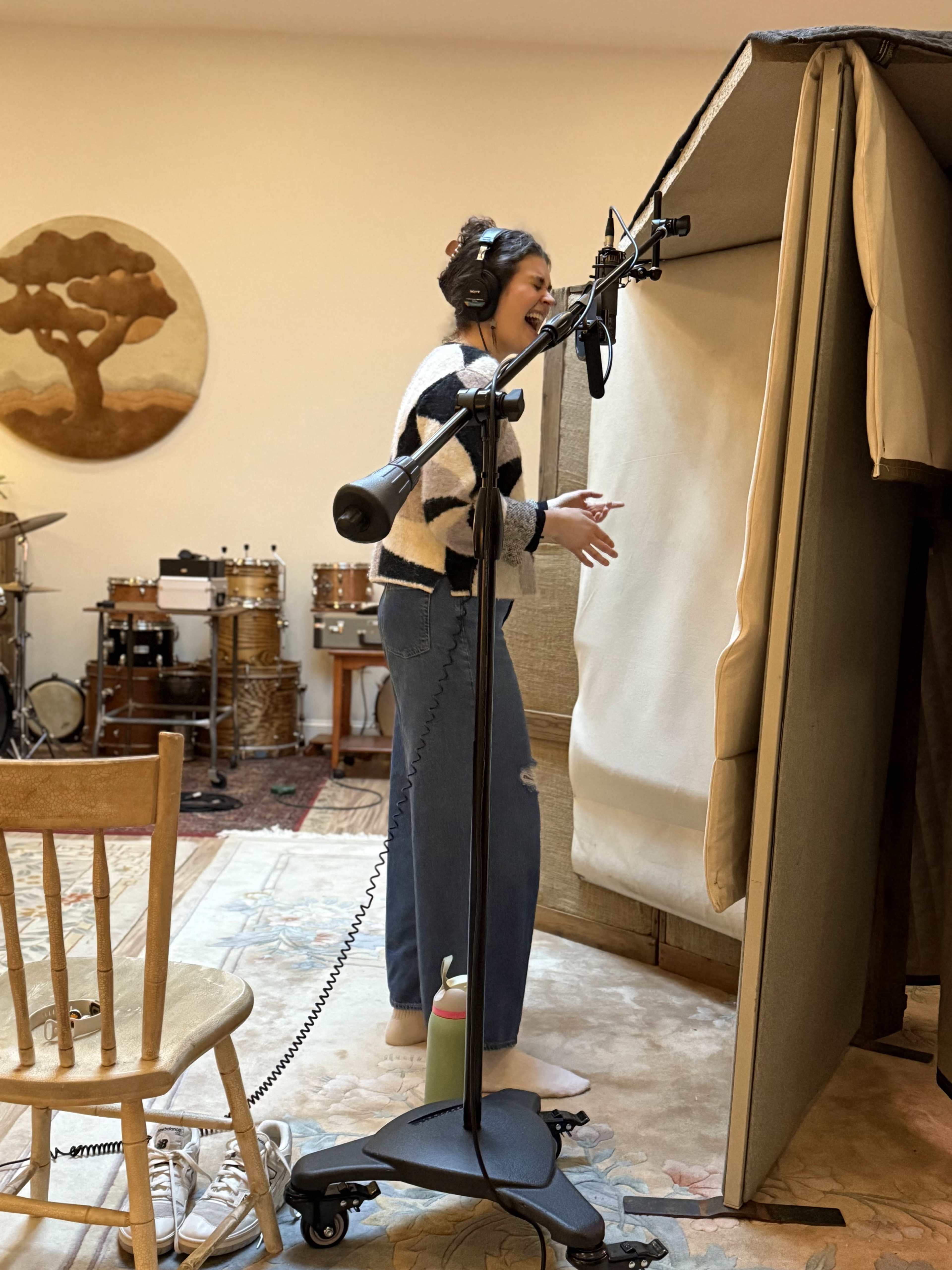 A person stands in a recording booth with a microphone, wearing headphones and casual clothing.