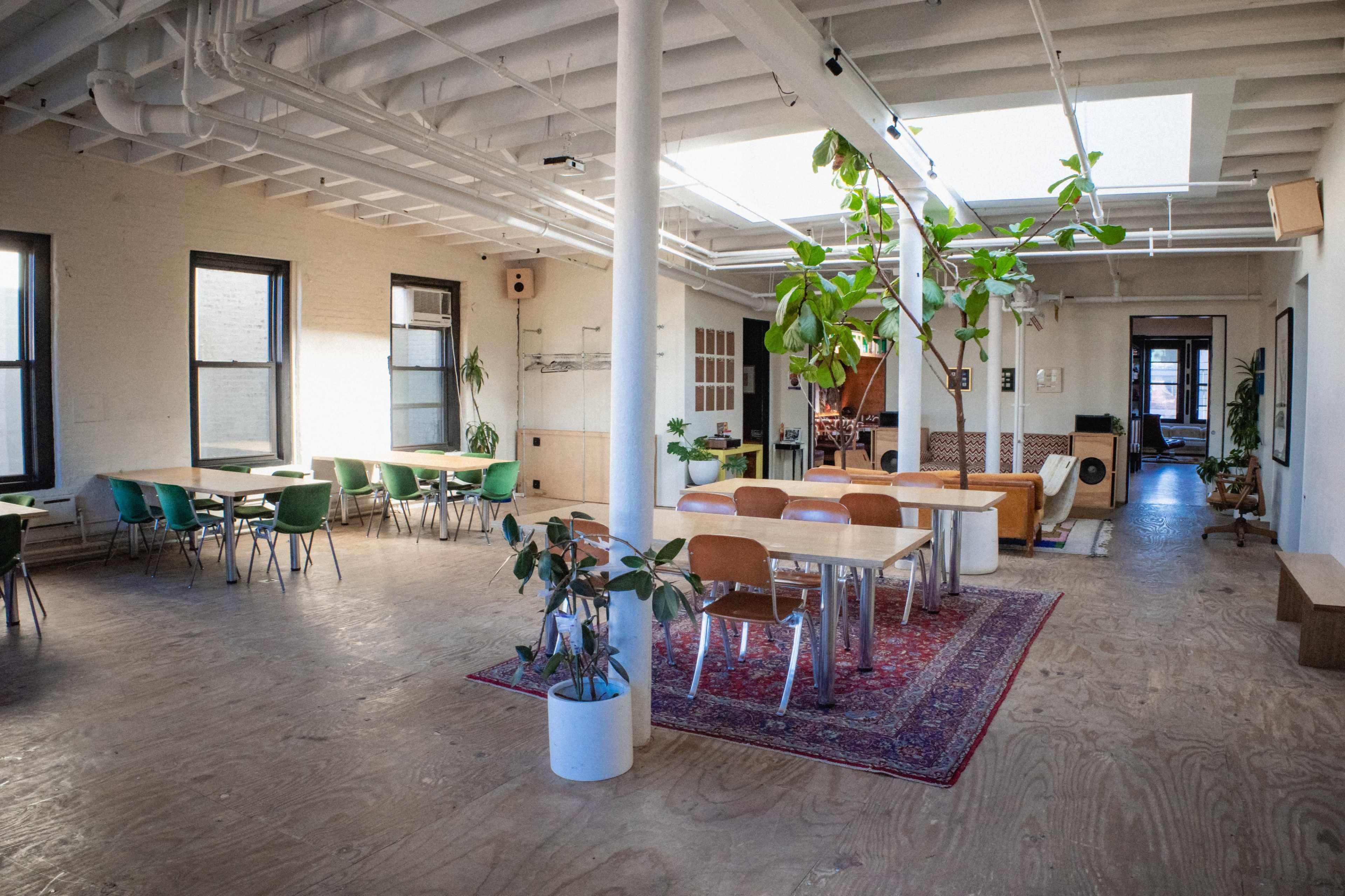 The image shows a spacious, modern communal area with wooden floors, green chairs, and plants, featuring tables for dining or work.