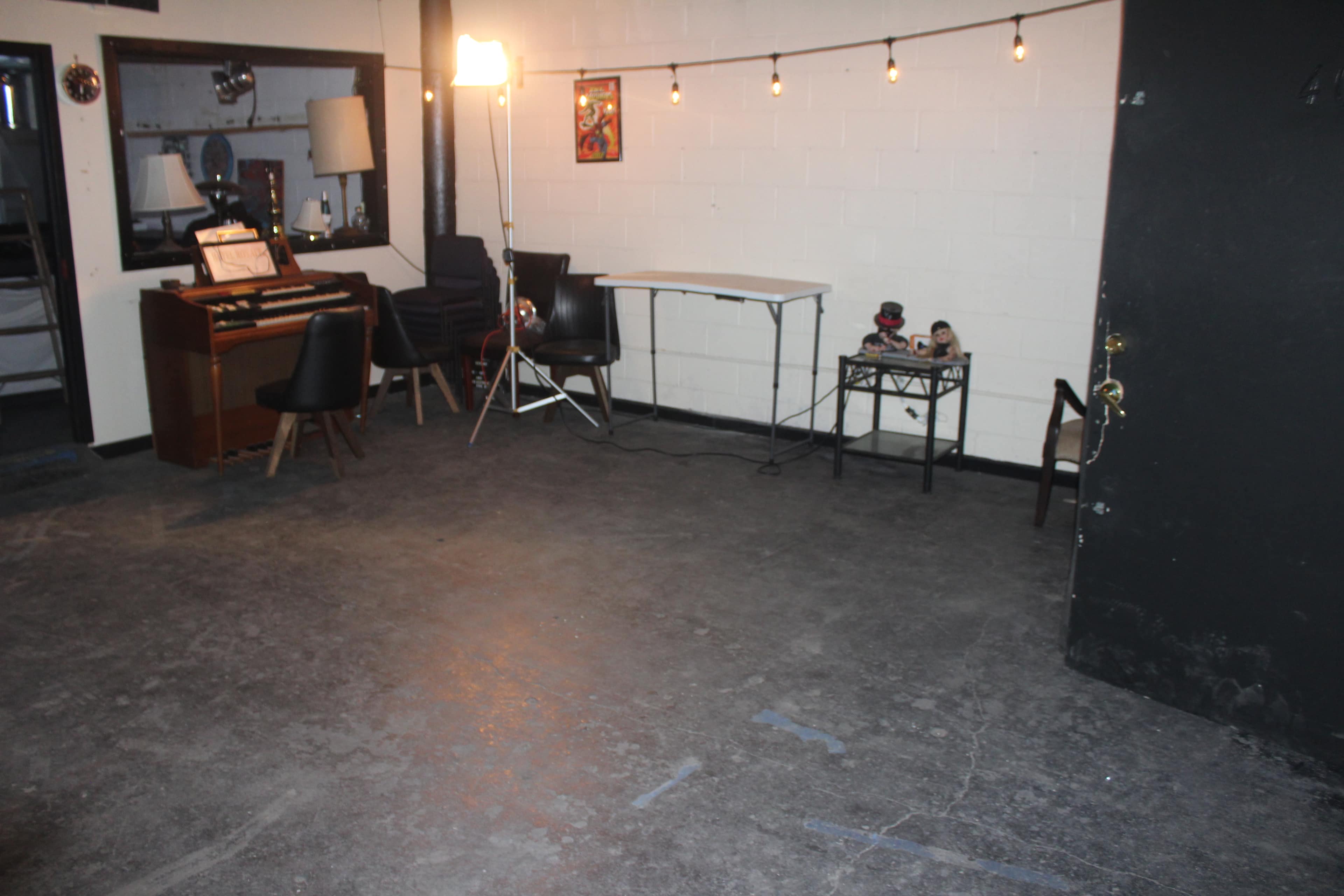 The image shows a sparsely furnished room with a piano, table, and chairs, illuminated by a standing light and featuring exposed string lights.
