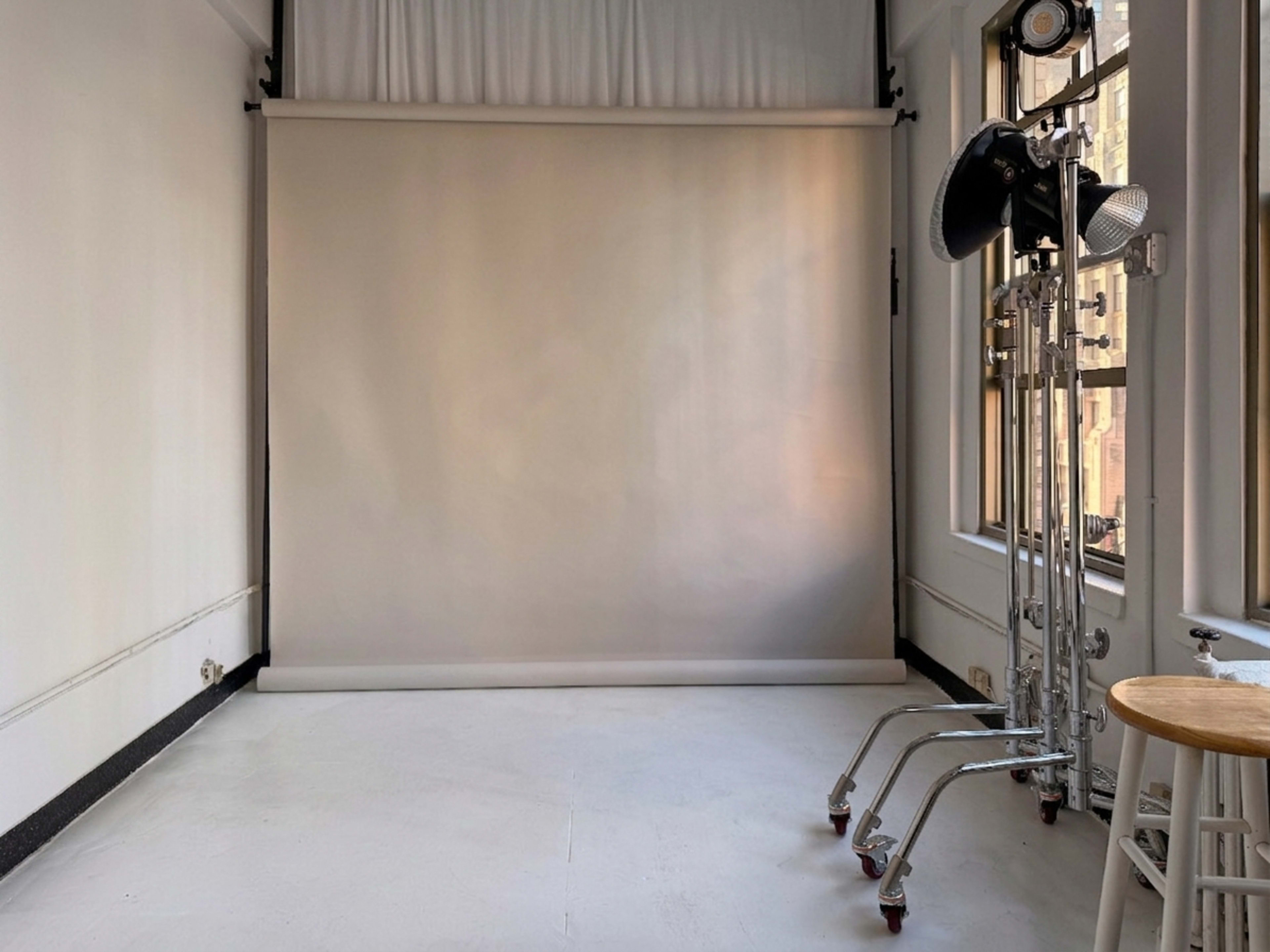 The image shows a minimalist photography studio featuring a gray backdrop and a stool, with studio lights on stands positioned to the side.