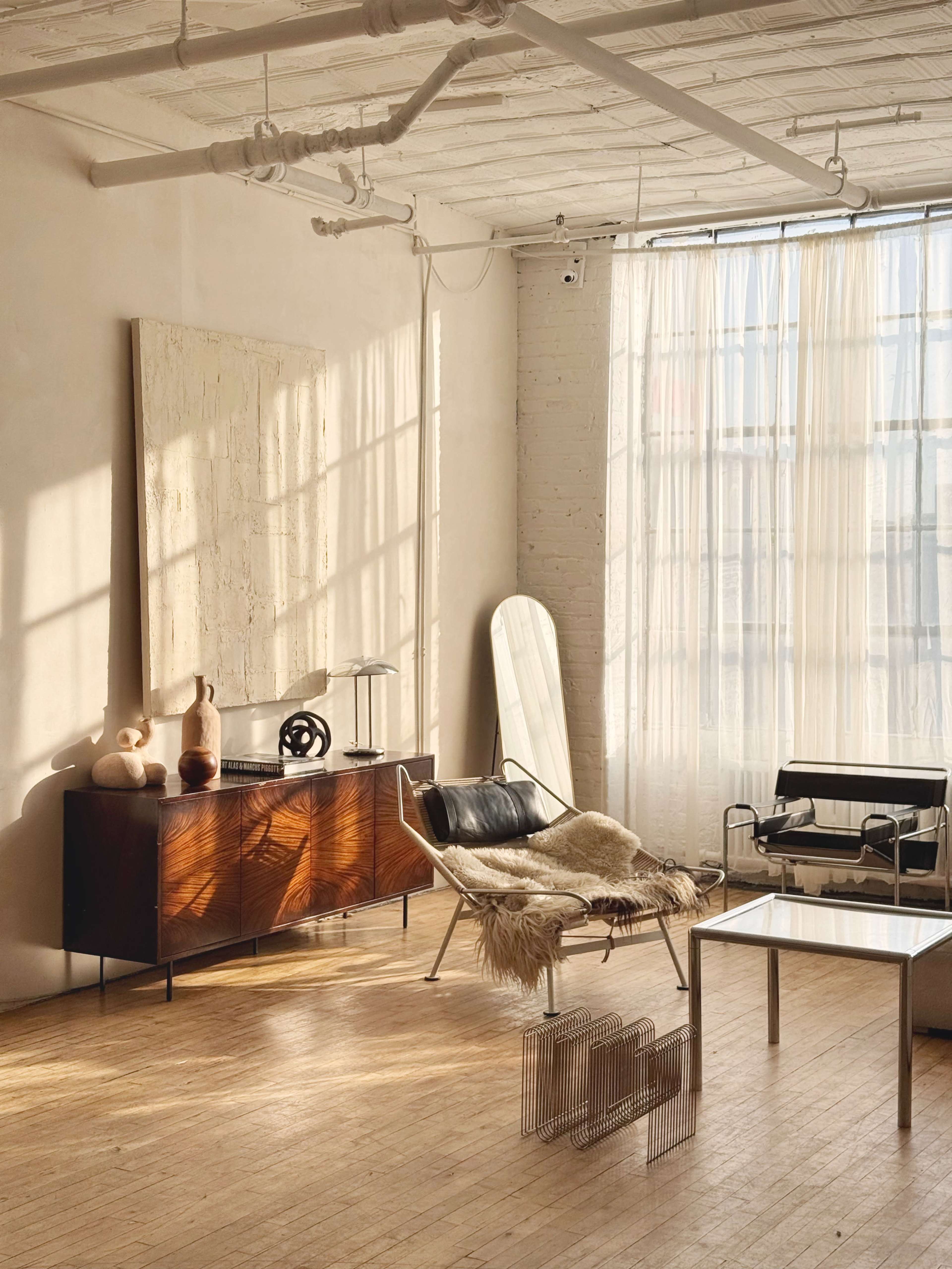 The image shows a modern, minimalistic living space with a leather lounge chair, a wooden sideboard, and large windows covered with sheer curtains.