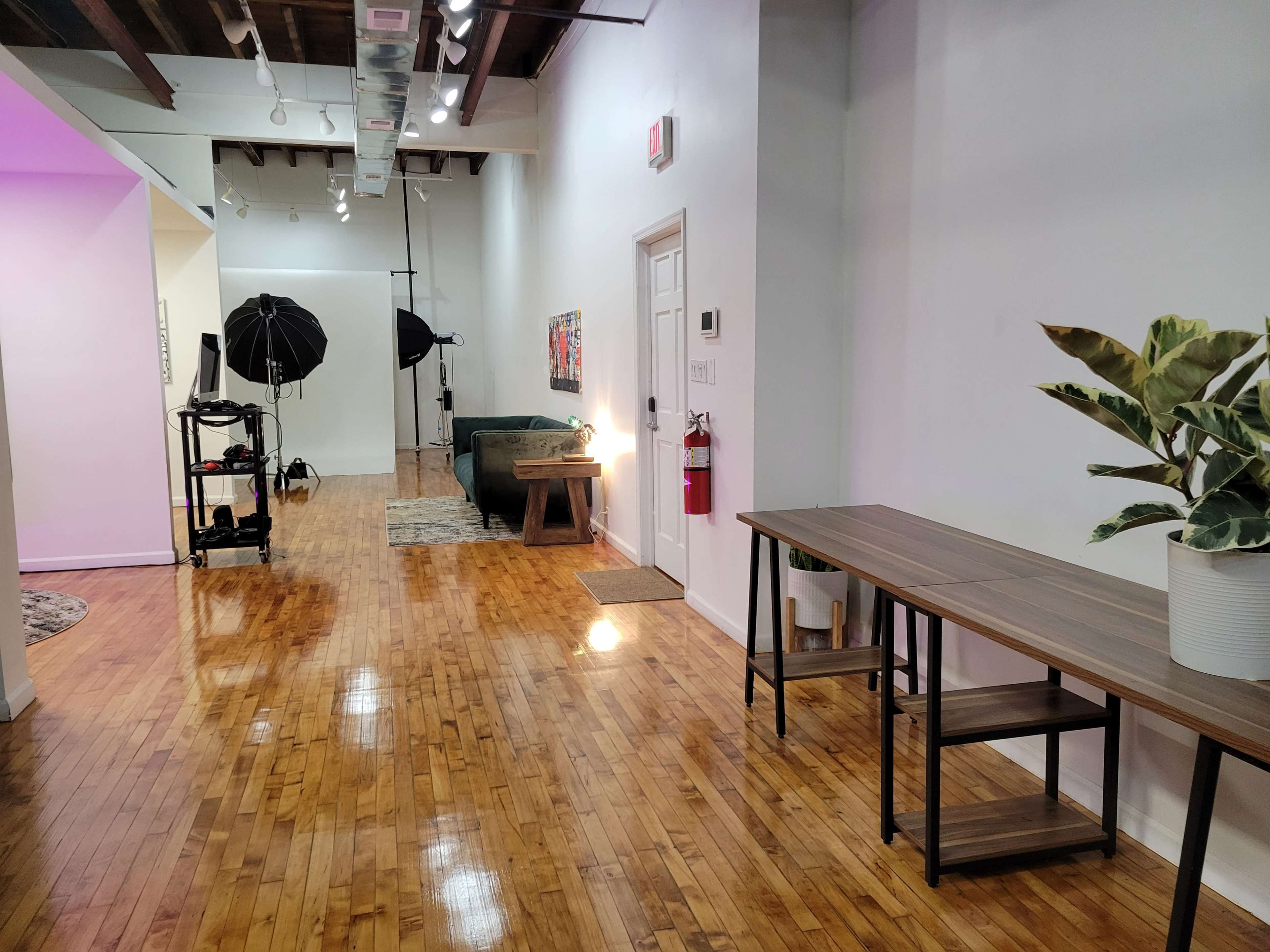 The image shows a spacious, brightly lit room with polished wooden floors, featuring a long table, a couch, photography equipment, and a potted plant.