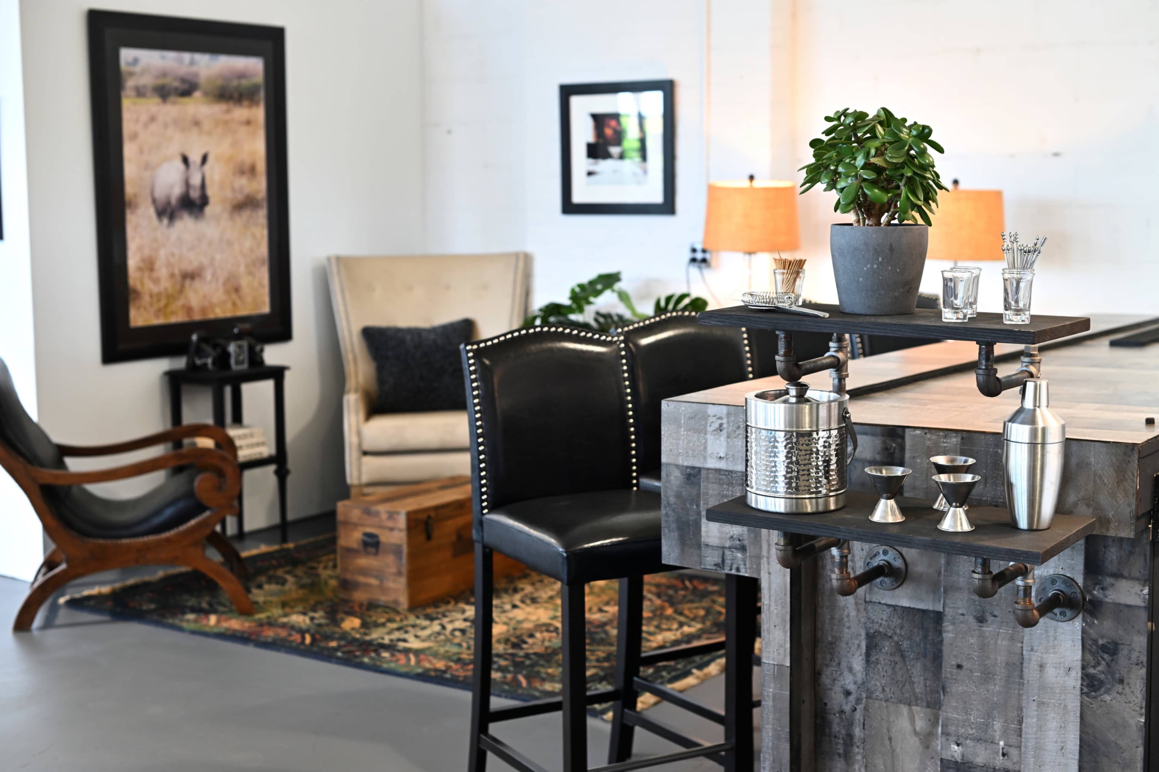 The image shows a stylish home interior with a wooden bar area featuring cocktail-making tools, black bar stools, and a cozy seating area with a chair and framed artwork on the wall.