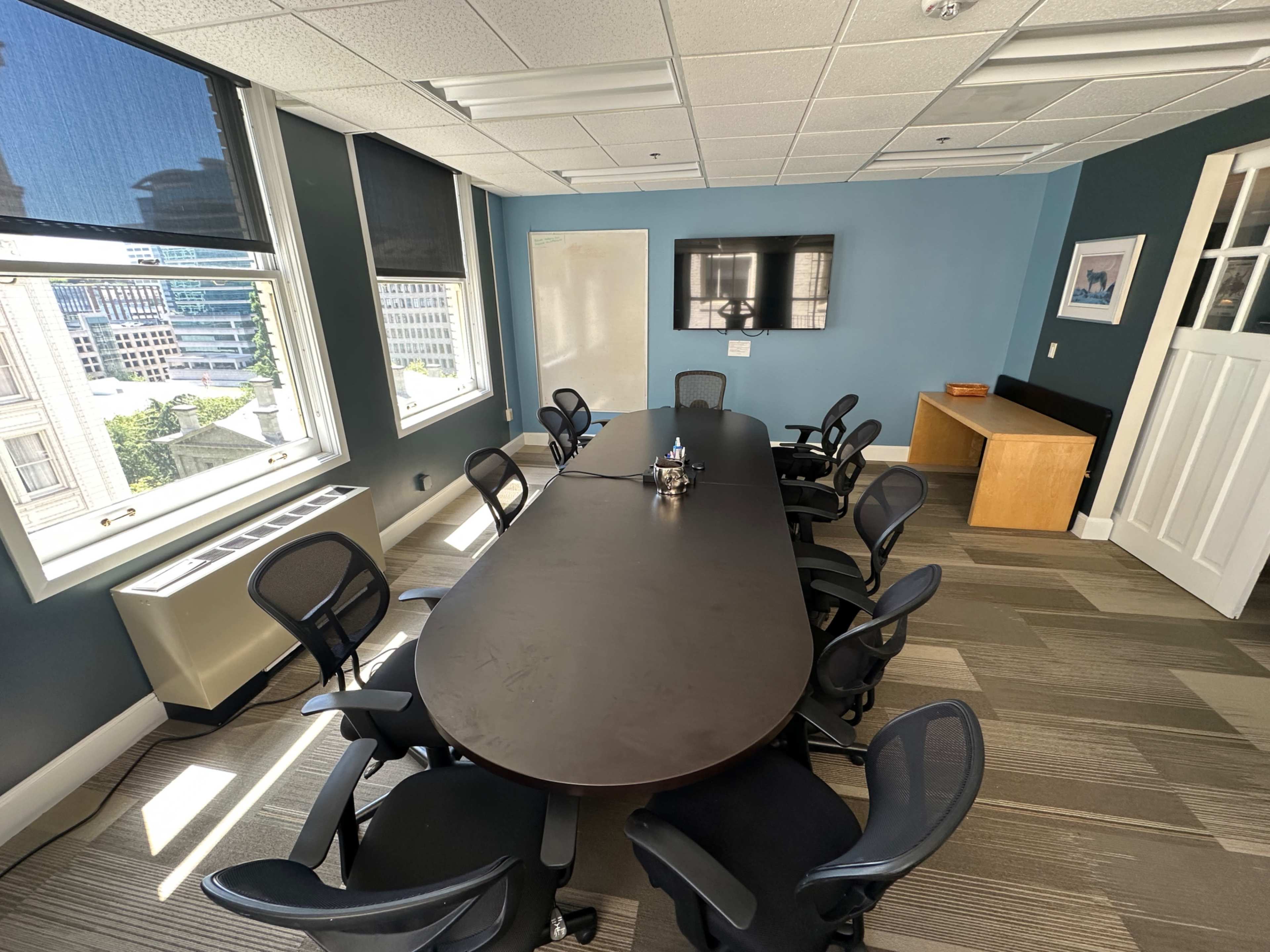 A modern conference room features a large oval table surrounded by ergonomic chairs, with a television mounted on the wall and windows providing natural light.
