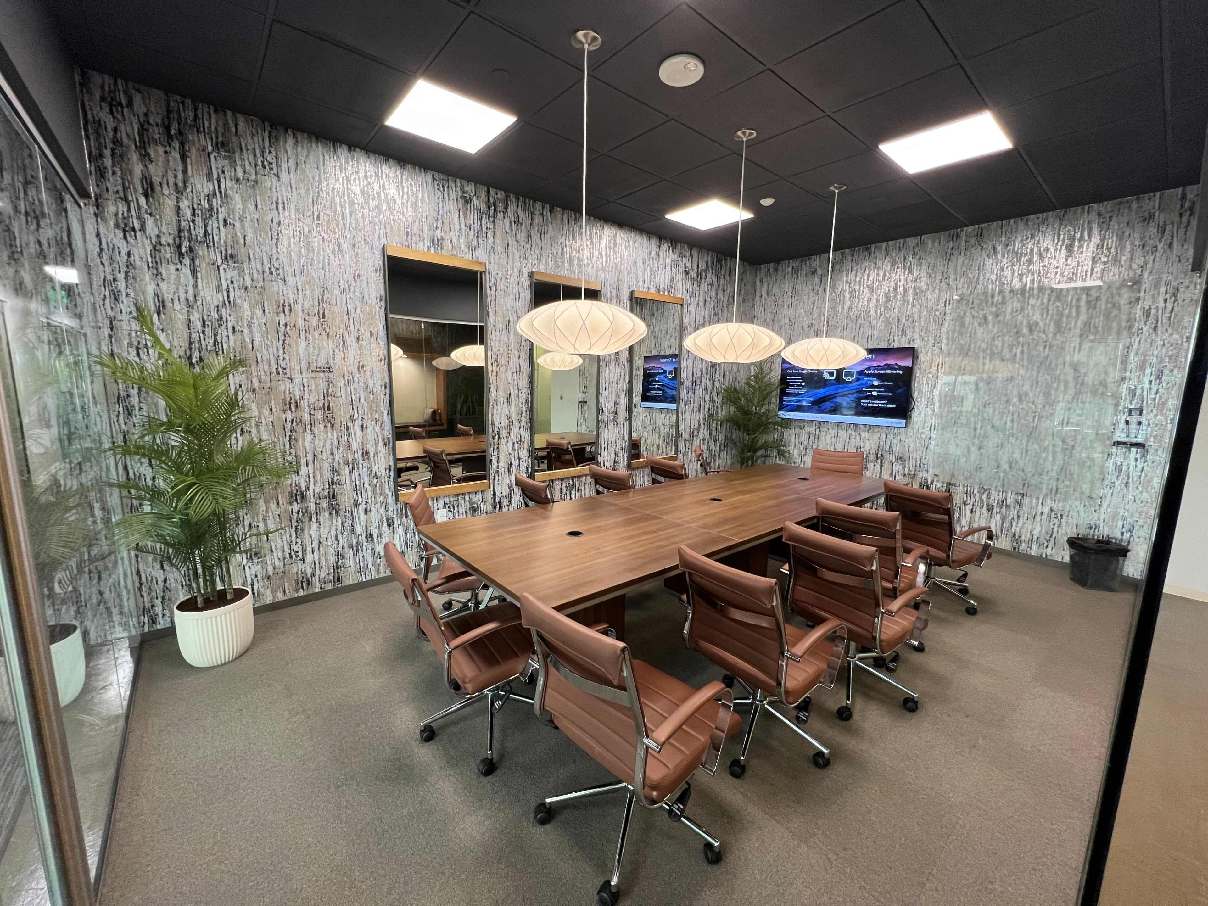 The image shows a modern conference room with a large wooden table surrounded by leather office chairs, illuminated by pendant lights, and features a textured wall and plants.