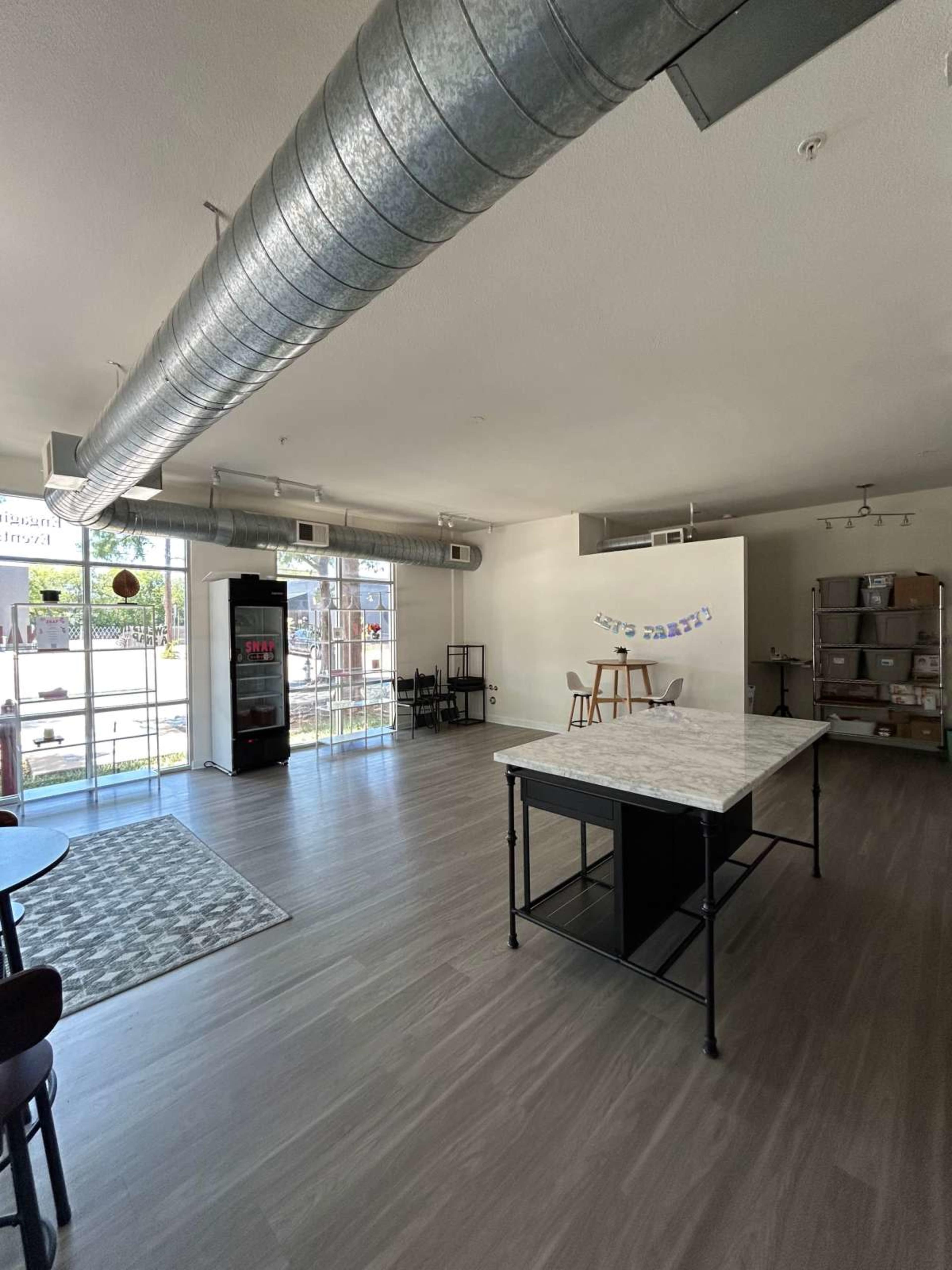 The image shows a spacious, modern interior of a café or community space with a kitchen island, seating area, and large windows.